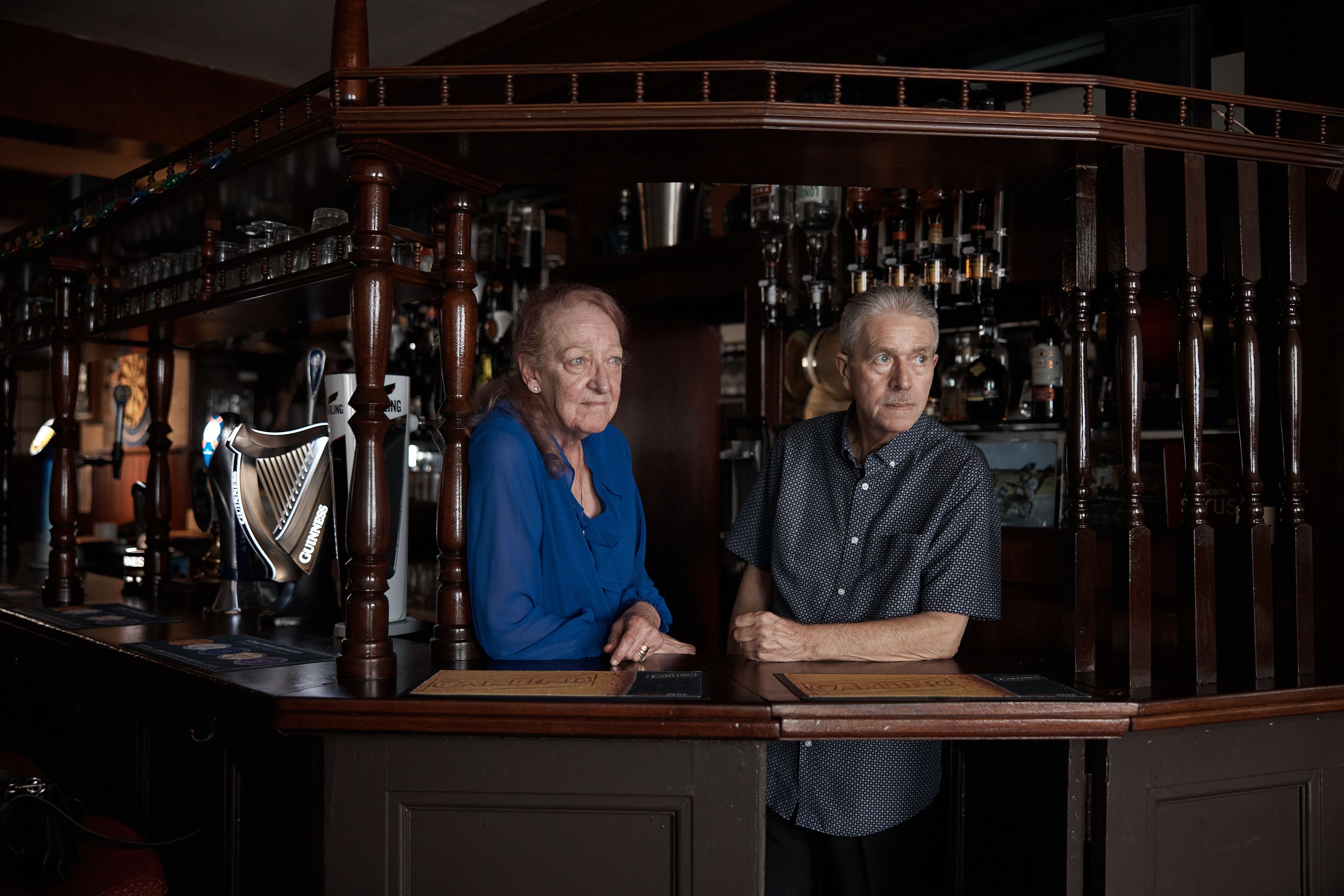 An older woman and man stand behind a bar with various beer taps, including one shaped like a harp.