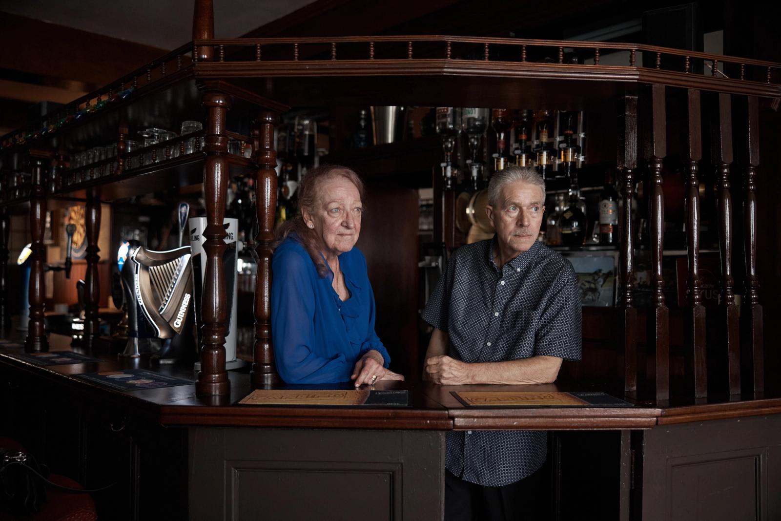 An older woman and man stand behind a bar with various beer taps, including one shaped like a harp.
