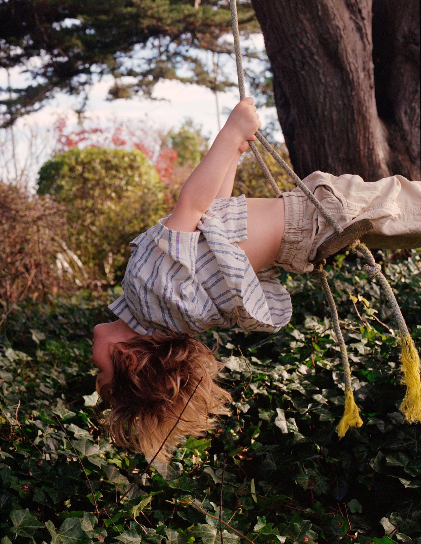 A young boy with messy brown hair is hanging upside down from a rope swing, wearing a striped shirt and khaki shorts, surrounded by green leaves and a tree trunk.
