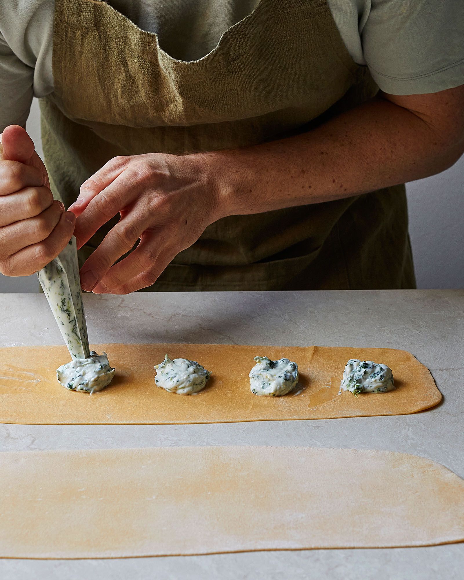 A person in a green apron is preparing a pasta dish, specifically filling pasta sheets with a mixture of cheese and herbs.