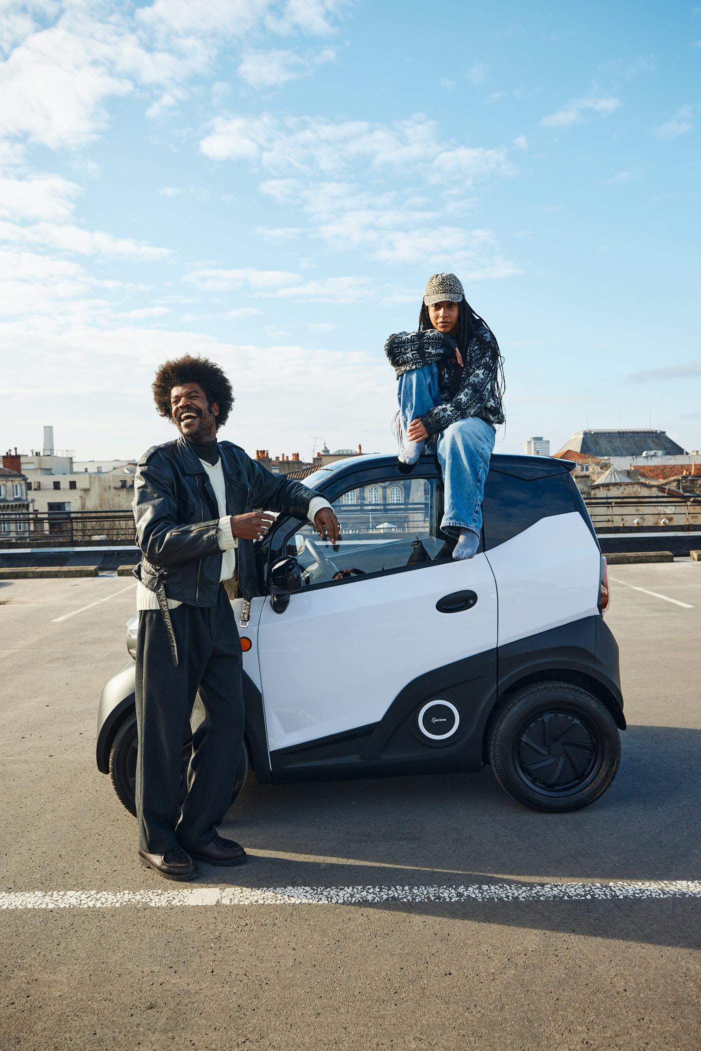 Two men, one with an Afro and one with dreadlocks, stand in a parking lot, one leaning on a car and the other sitting on top of it. The atmosphere is casual and relaxed, with a.