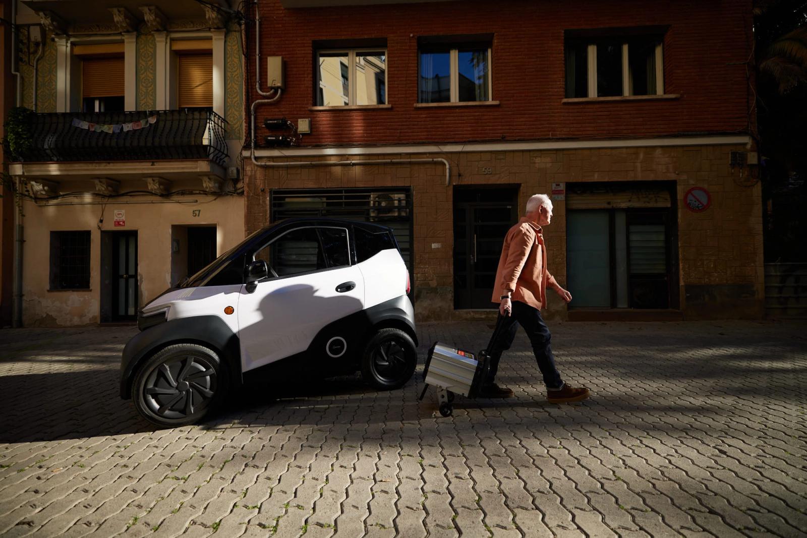 An older man with a white buzz cut and a warm orange shirt walks down a cobblestone street, carrying a small white car on a wheeled dolly. He is pushing it with his right hand,.
