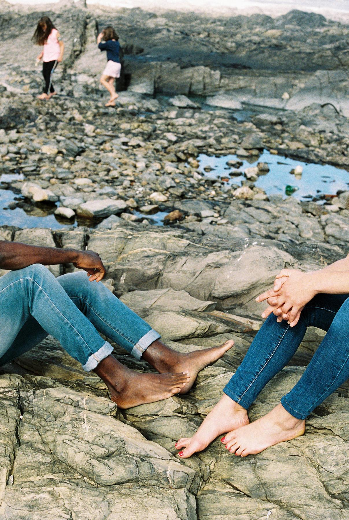 Two people, one with dark skin and one with light skin, sitting on a rocky beach, both barefoot and wearing blue jeans.