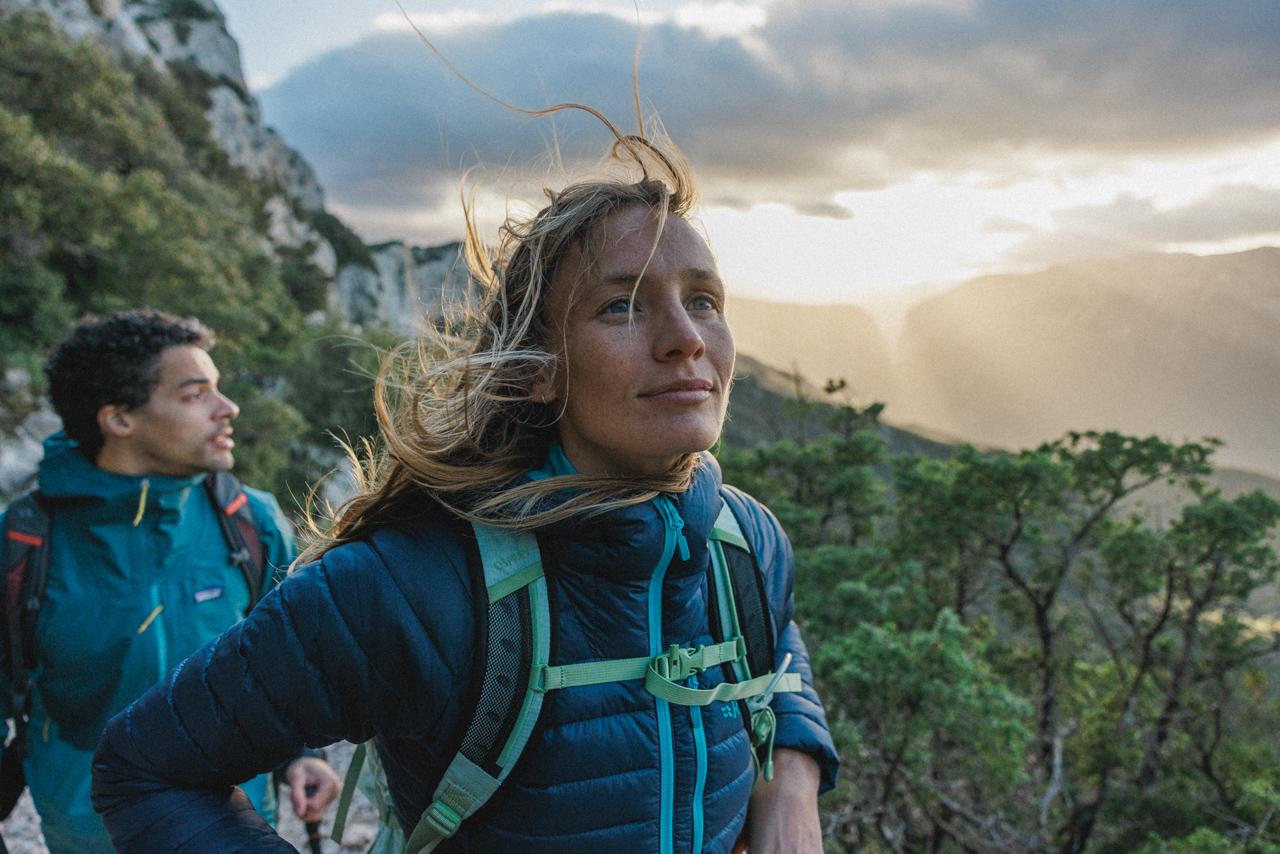 A woman with long blonde hair and a blue jacket hiking in a mountainous landscape, with a man in the background wearing a similar blue jacket.