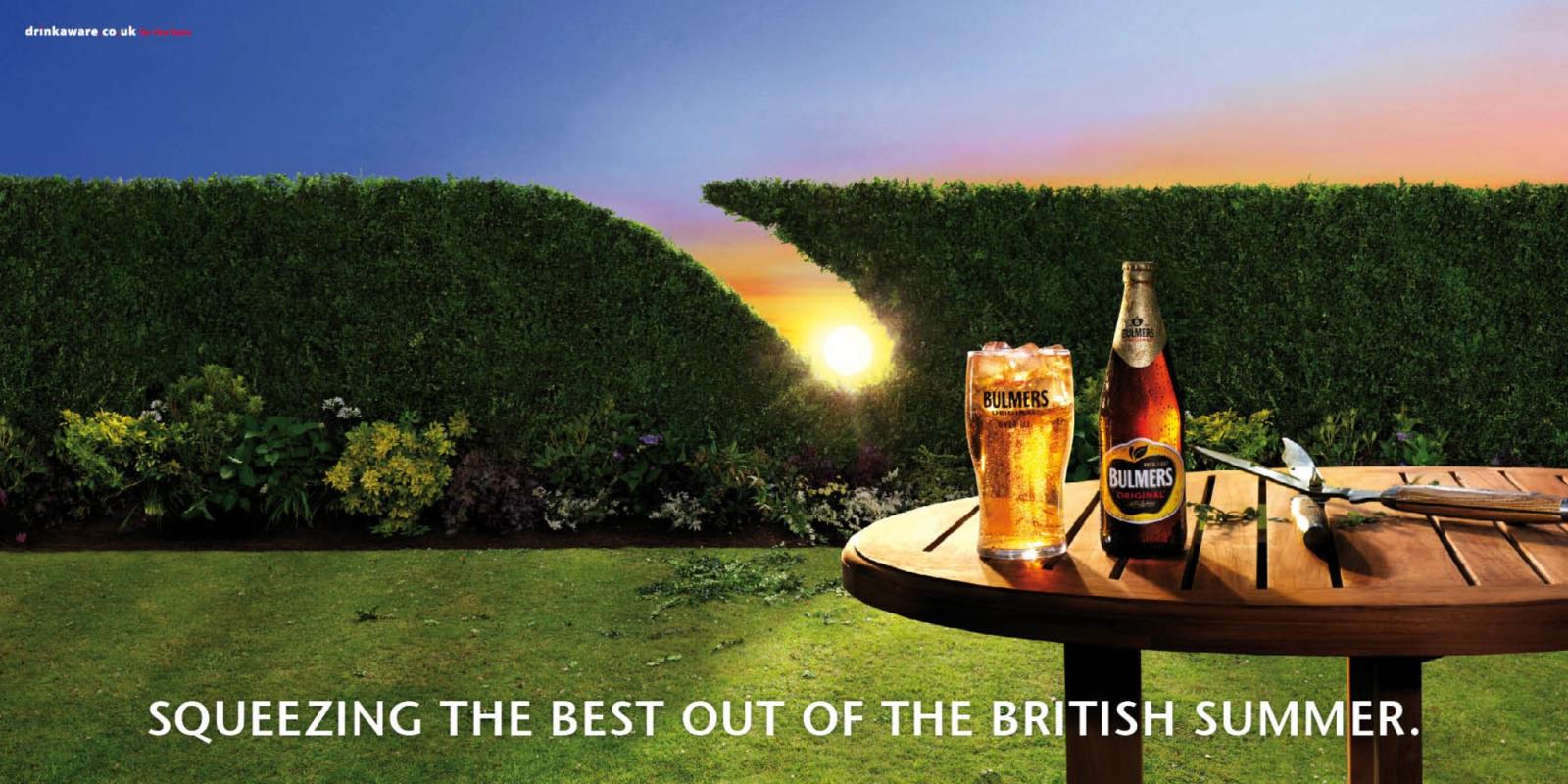 A person is sitting on a grassy lawn, holding a glass of beer and a plate of food, with a beautiful sunset in the background.