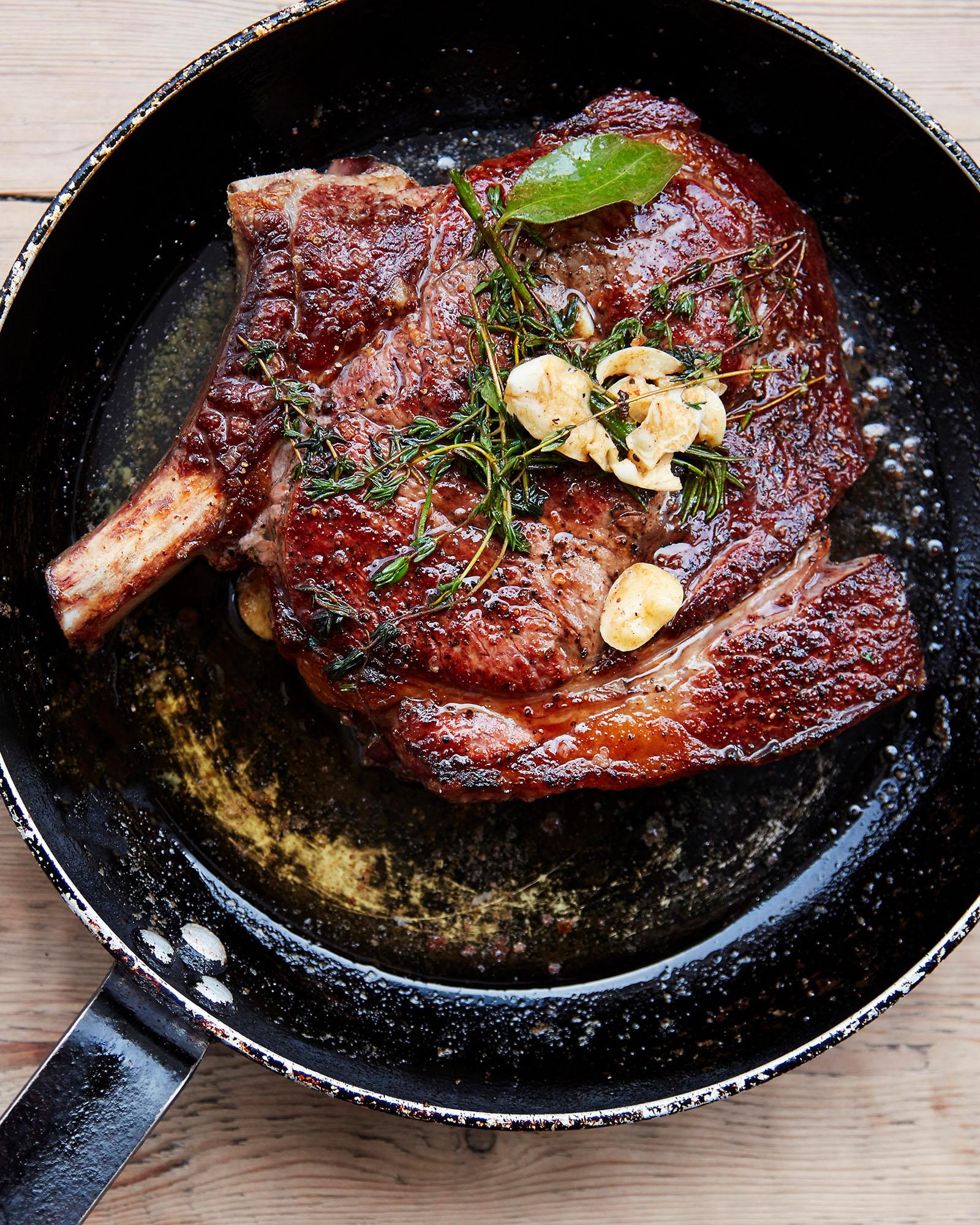 A pan containing a cooked steak with herbs and garlic on top, sitting on a wooden table.