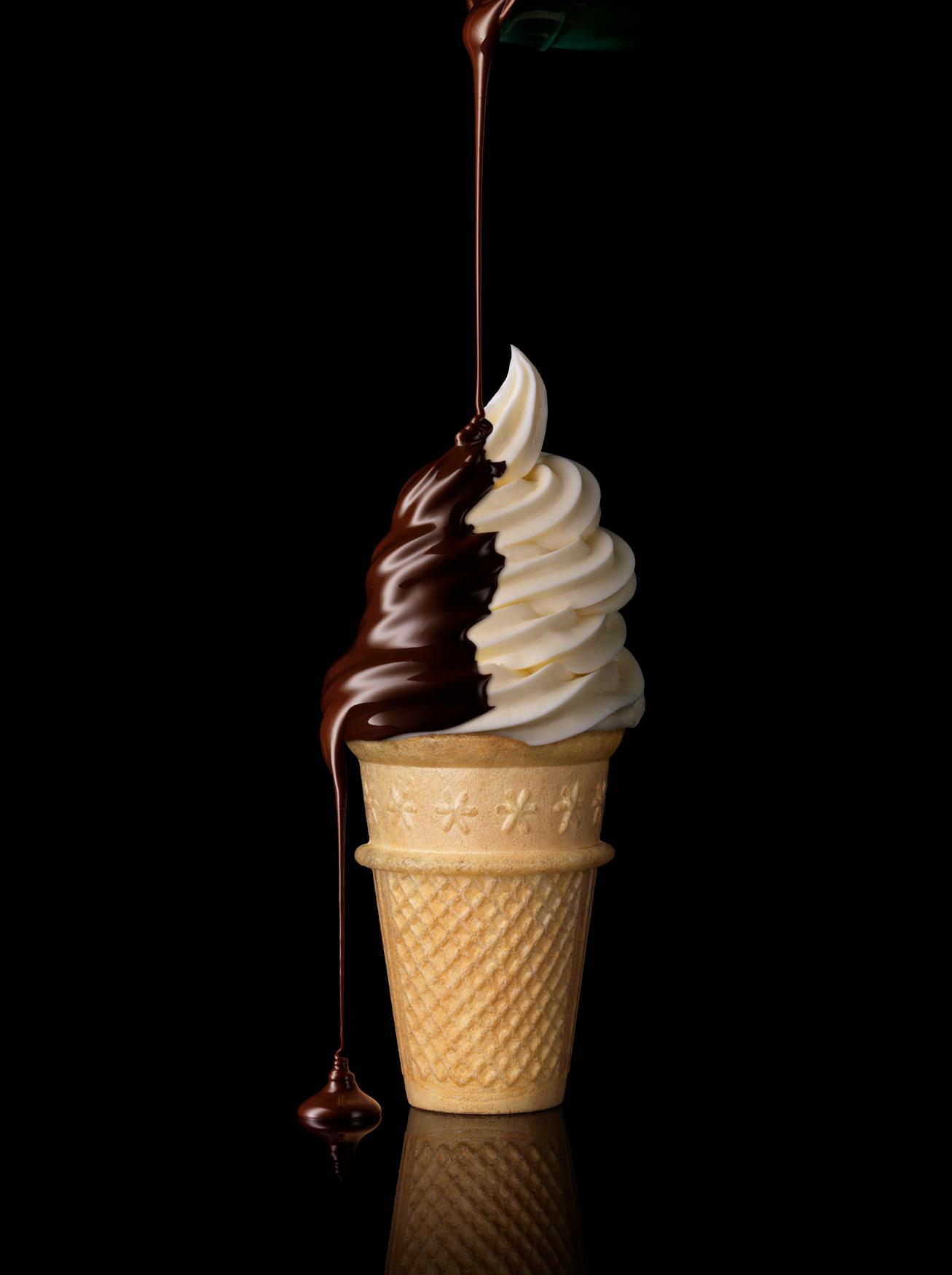A chocolate ice cream cone with a generous helping of chocolate sauce being poured over the top.