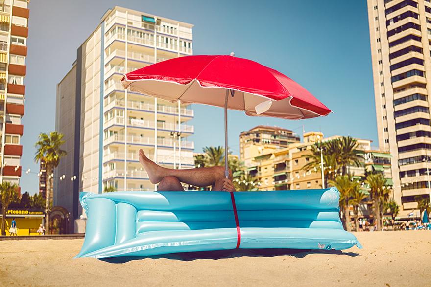 A person lies on a beach under an umbrella near a large blue inflatable structure with a red strap, surrounded by palm trees and high-rise buildings.