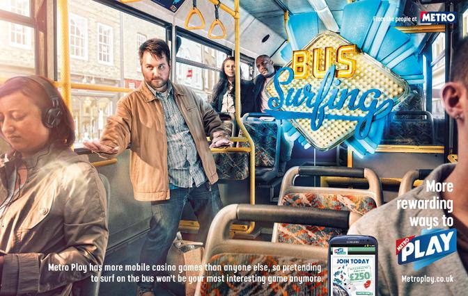 A man on a bus, wearing a brown jacket and jeans, looking at his phone, with a woman sitting next to him wearing headphones, and a man standing in the background looking at the camera.