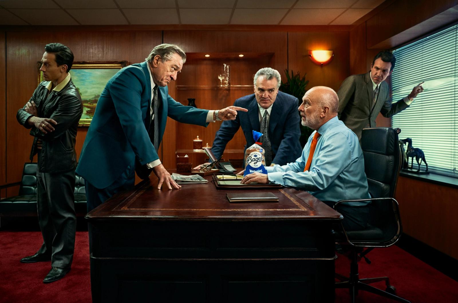 A group of men in business attire, including a suit and tie, are gathered around a desk, with a bag of Bage's bread on the table, in a modern office setting.