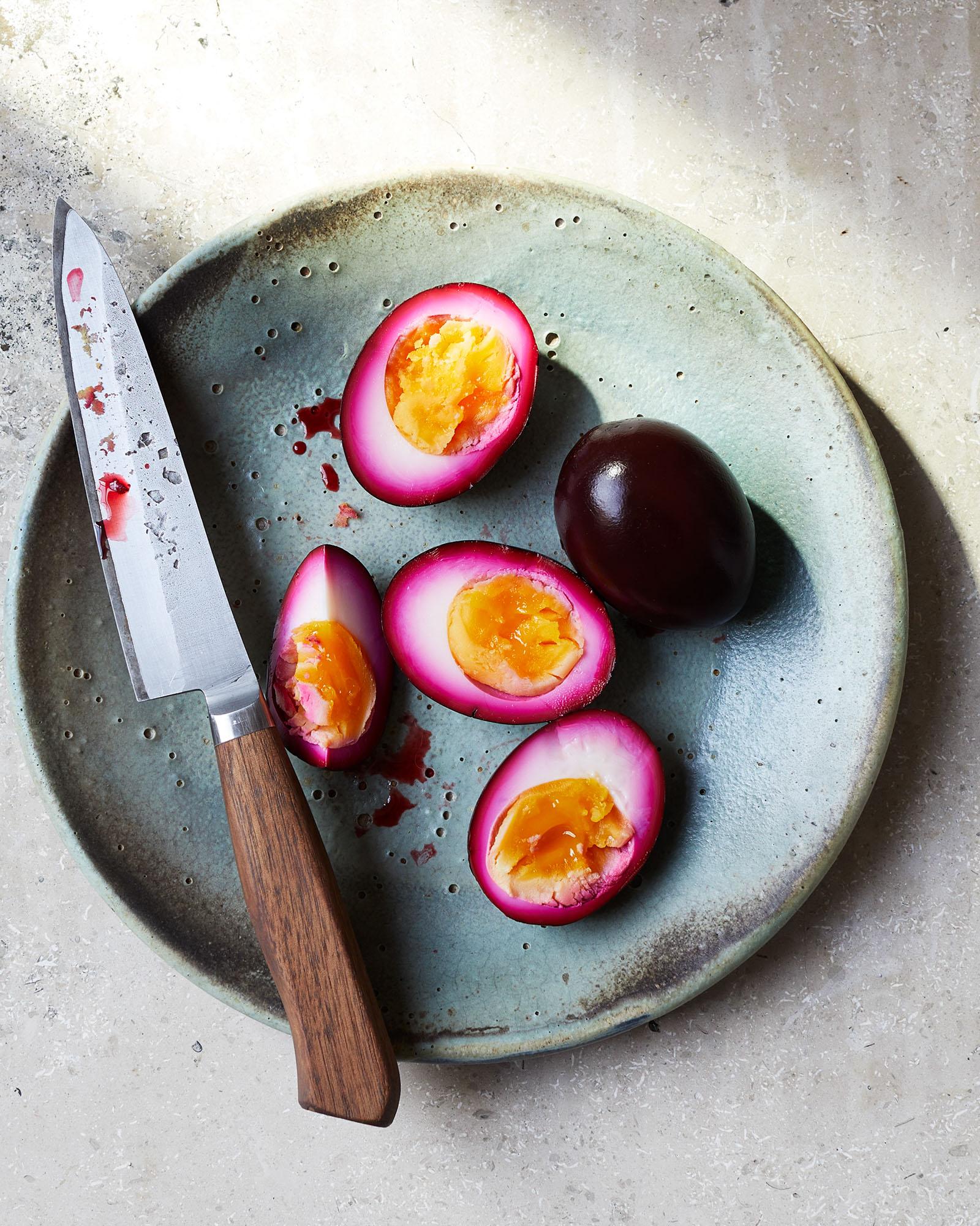 A plate of pickled eggs, with a few eggs sliced in half to show the pickled yellow centres, and a small knife with a wooden handle lying across the plate.