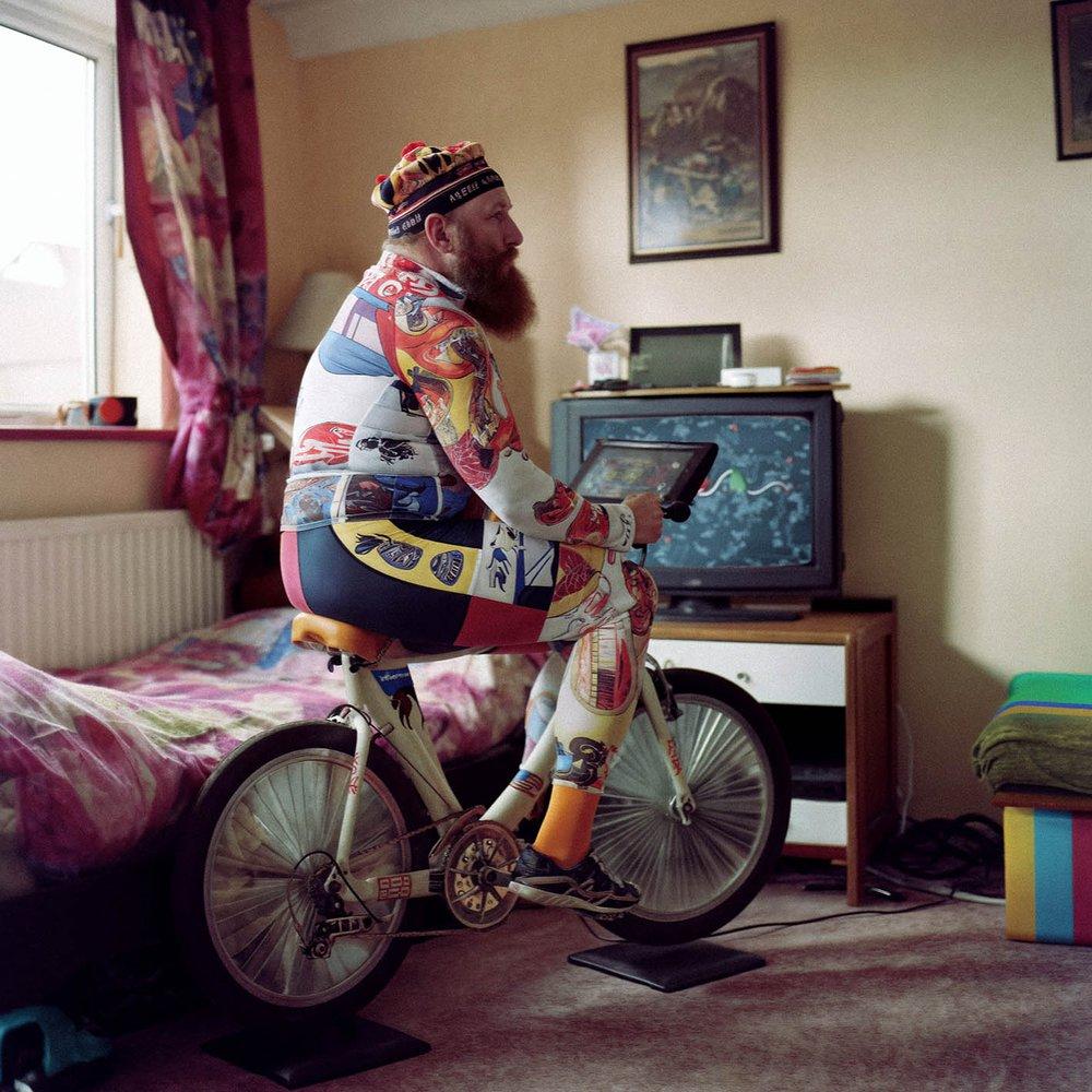A man wearing a colorful, patterned onesie is sitting on a bicycle in a bedroom, surrounded by a bed, a TV, and a window.