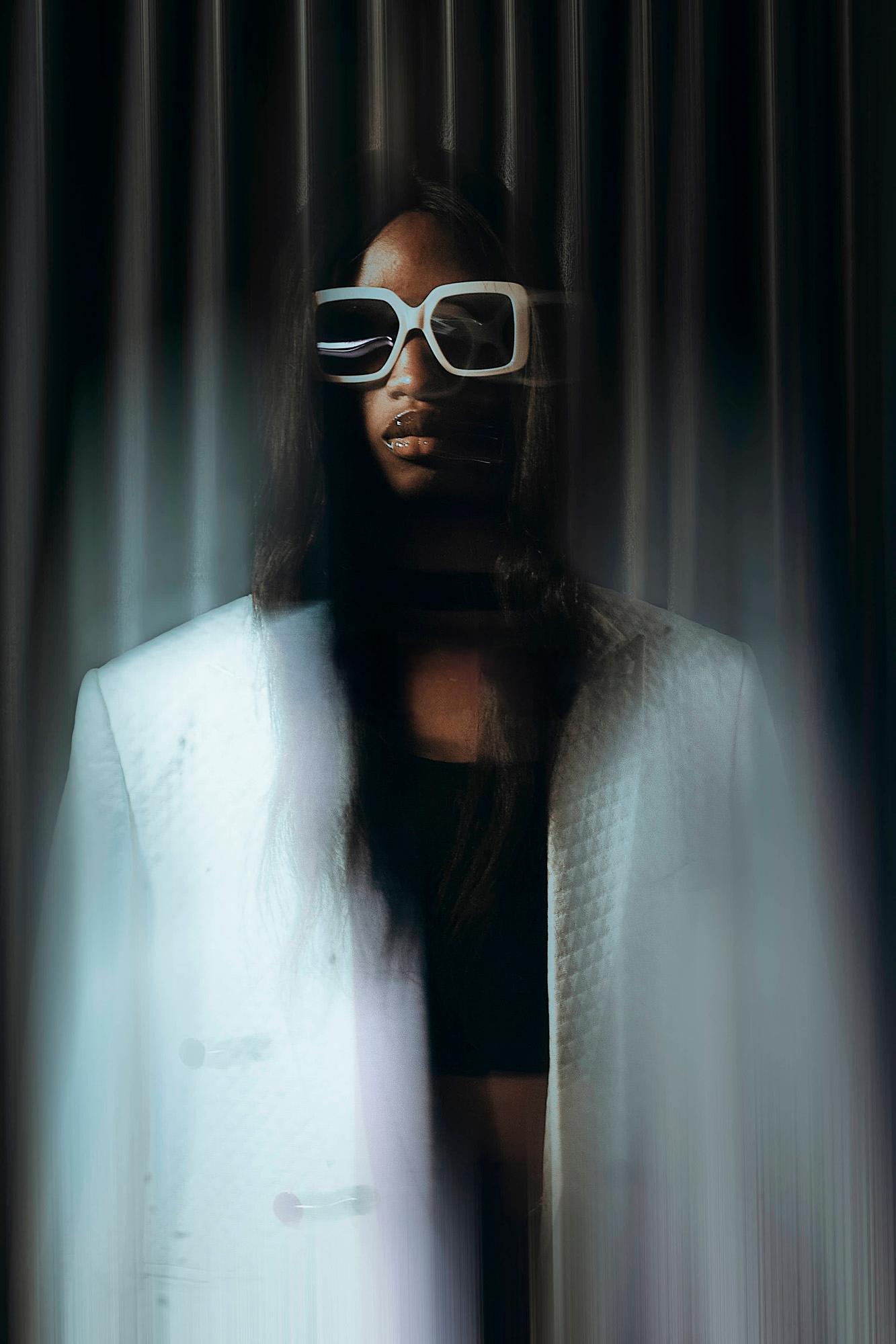 A woman wearing a white jacket and sunglasses, standing in front of a dark background.