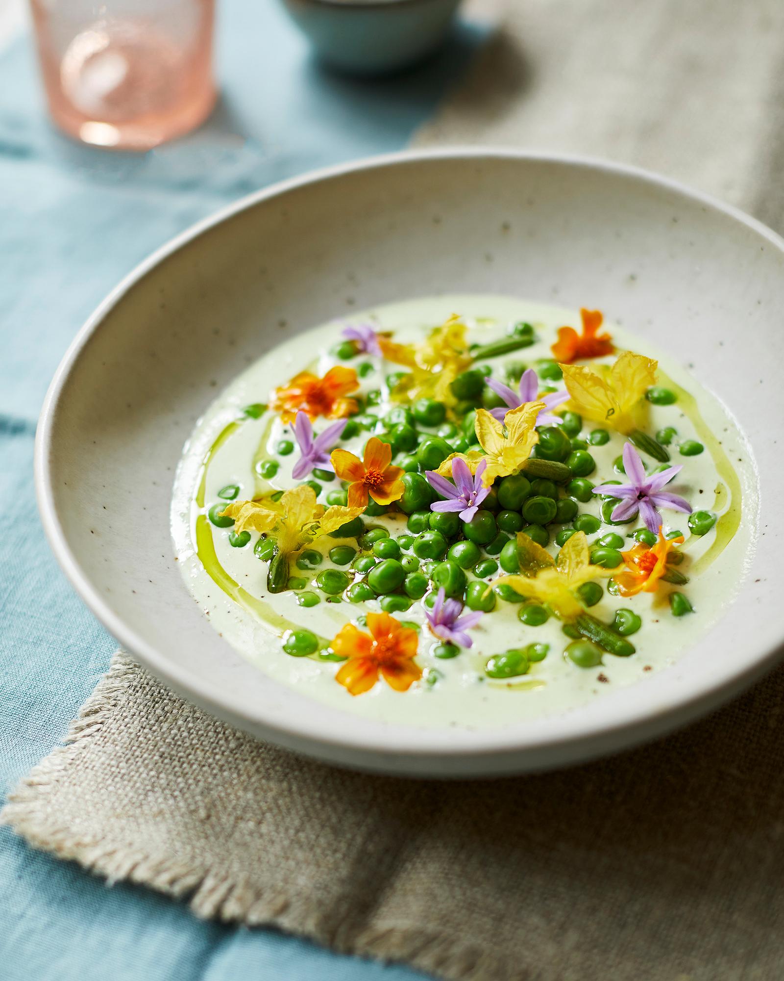 A bowl of creamy soup with a variety of vegetables and a few edible flowers on top, served in a white ceramic bowl on a light blue tablecloth.