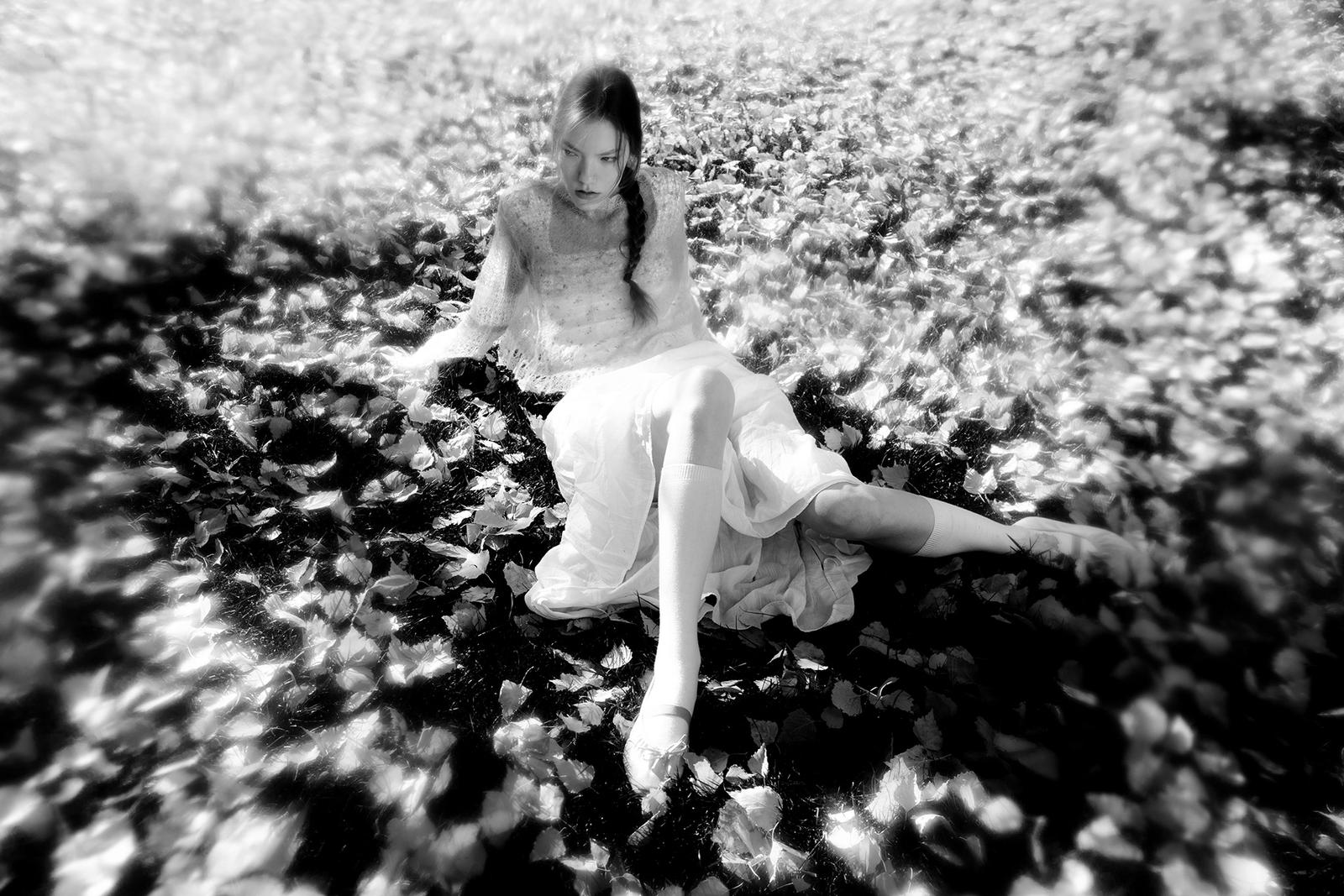 A woman in a white wedding dress and high heels is sitting on a bed of leaves, surrounded by a forest of trees.
