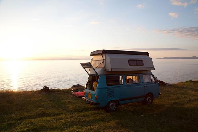 A blue and white VW camper van is parked on a cliff overlooking the ocean at sunset.