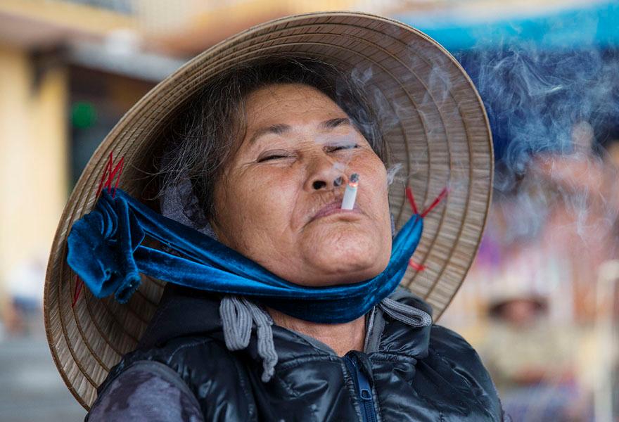 A woman wearing a traditional Vietnamese conical hat, smoking a cigarette, with a blurred background.