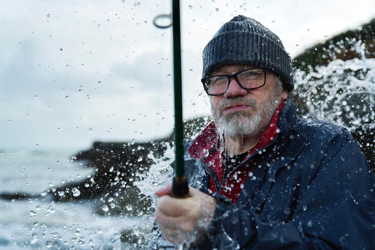 A man in a wetsuit and a beanie is holding a fishing rod in the ocean, surrounded by water splashes.