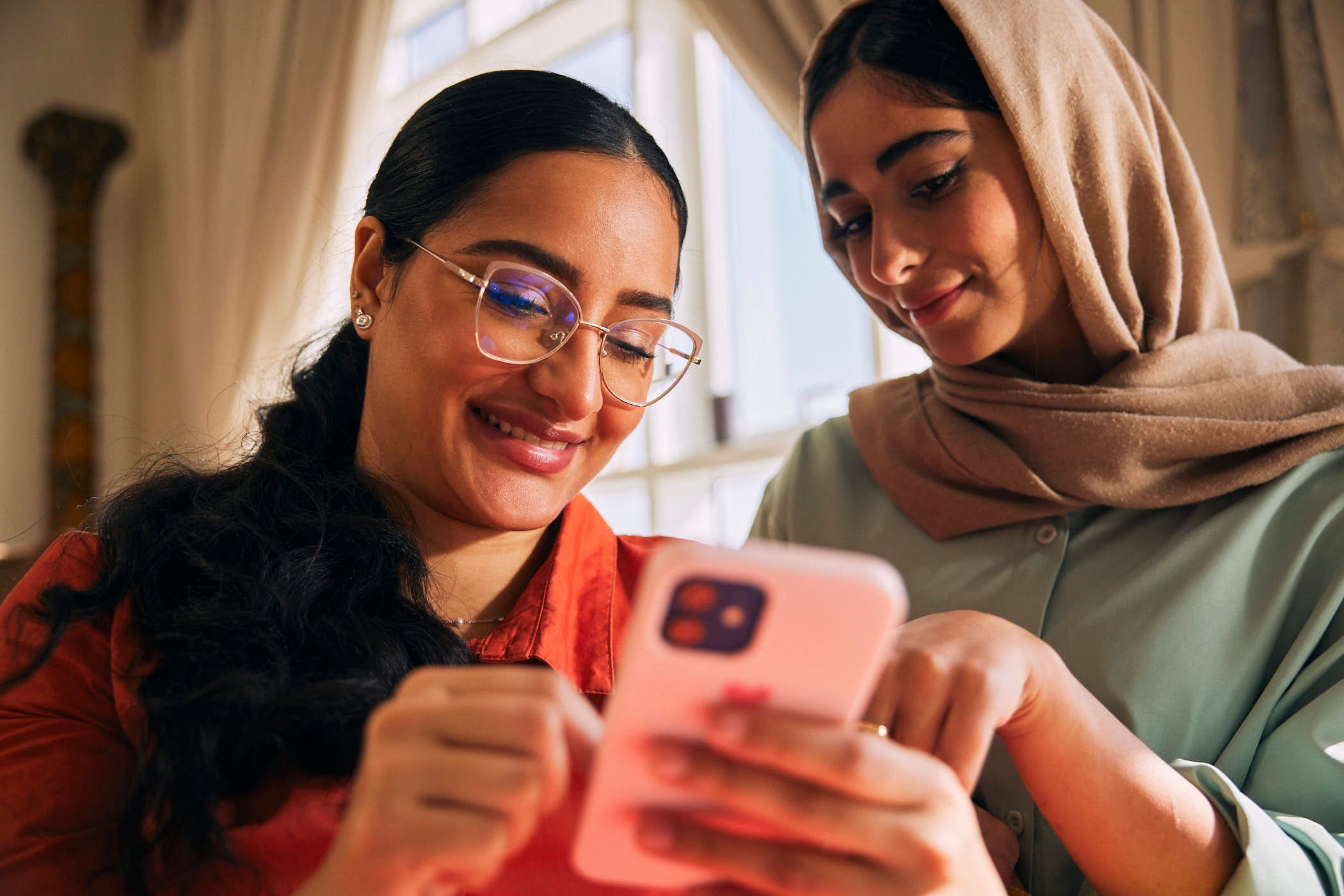 Two women, one wearing a hijab, look at a phone together, smiling and laughing. They appear to be sharing a funny or interesting moment.