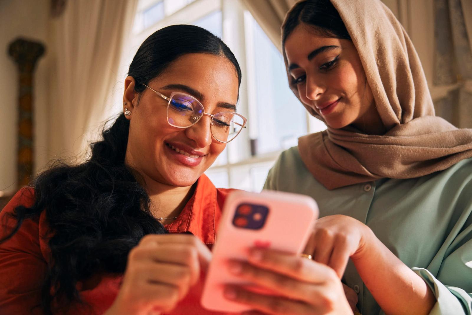 Two women, one wearing a hijab, look at a phone together, smiling and laughing. They appear to be sharing a funny or interesting moment.