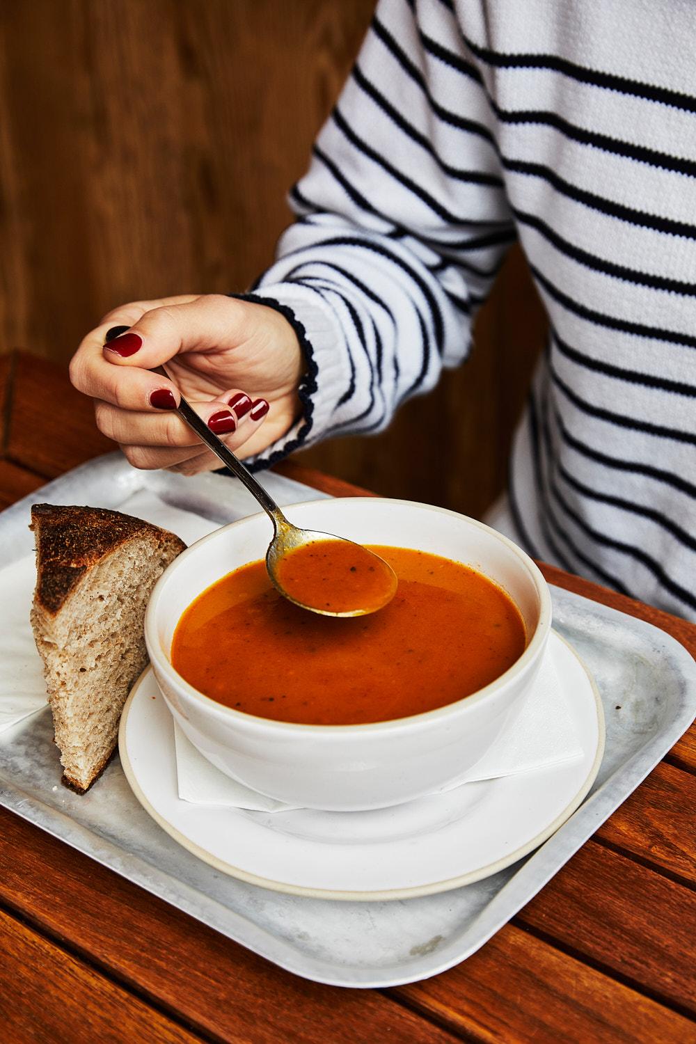 A woman's hand is holding a spoon above a bowl of tomato soup, with a slice of bread on the side.