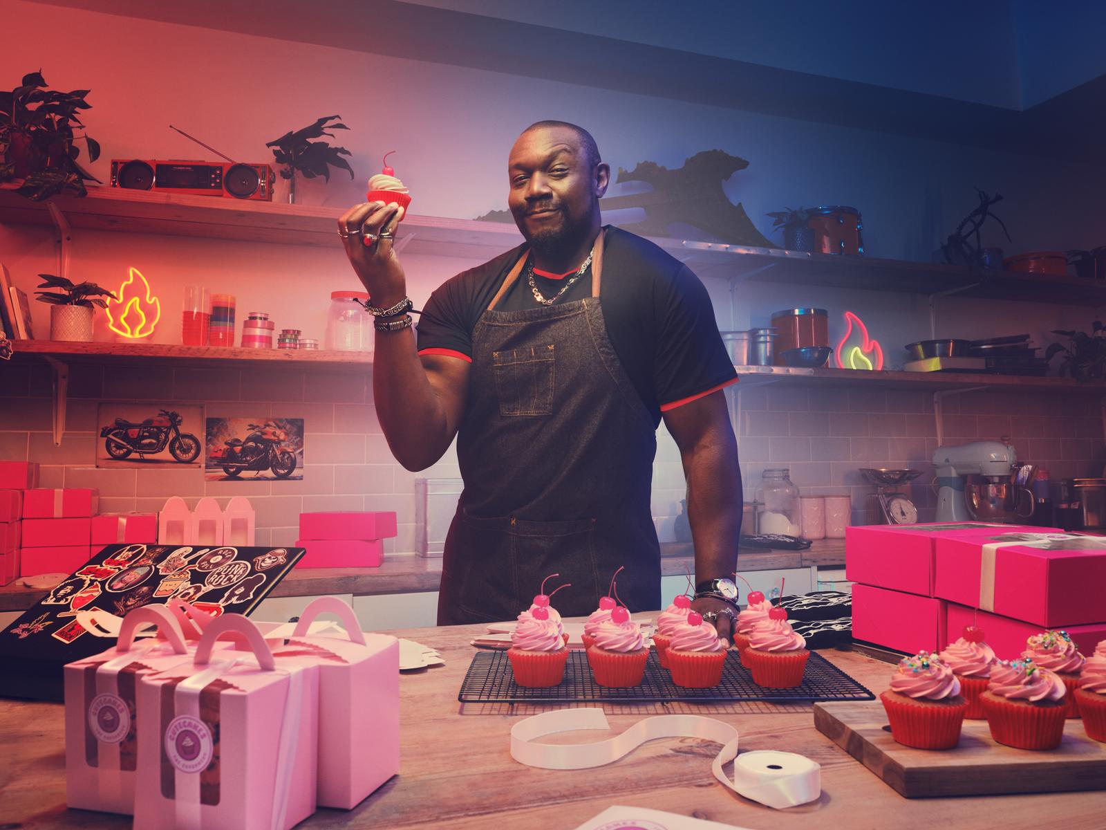 A man in a black apron and chef's hat, holding a cupcake with pink frosting and a cherry on top, in a kitchen with a counter and shelves with baking supplies.