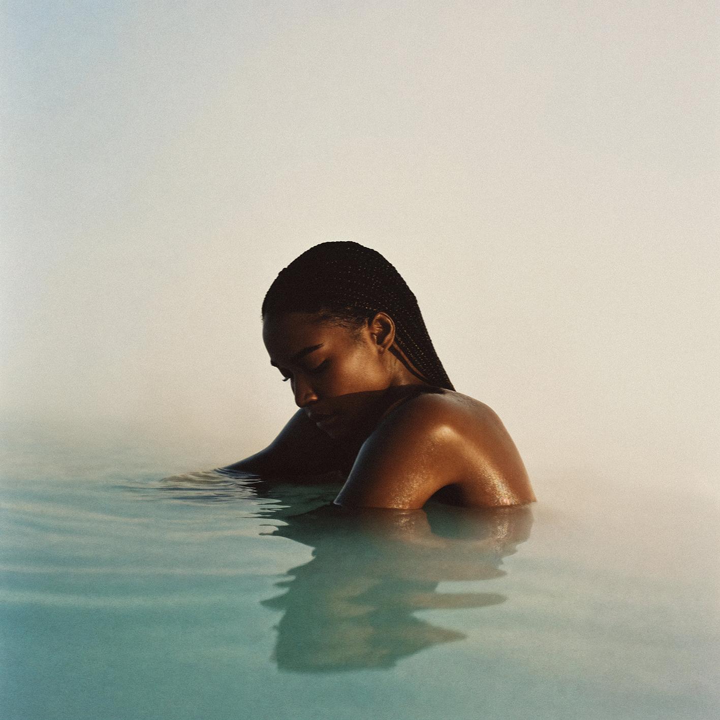 A woman with dark skin and braided hair is submerged in water, her face tilted upwards, with a subtle expression.