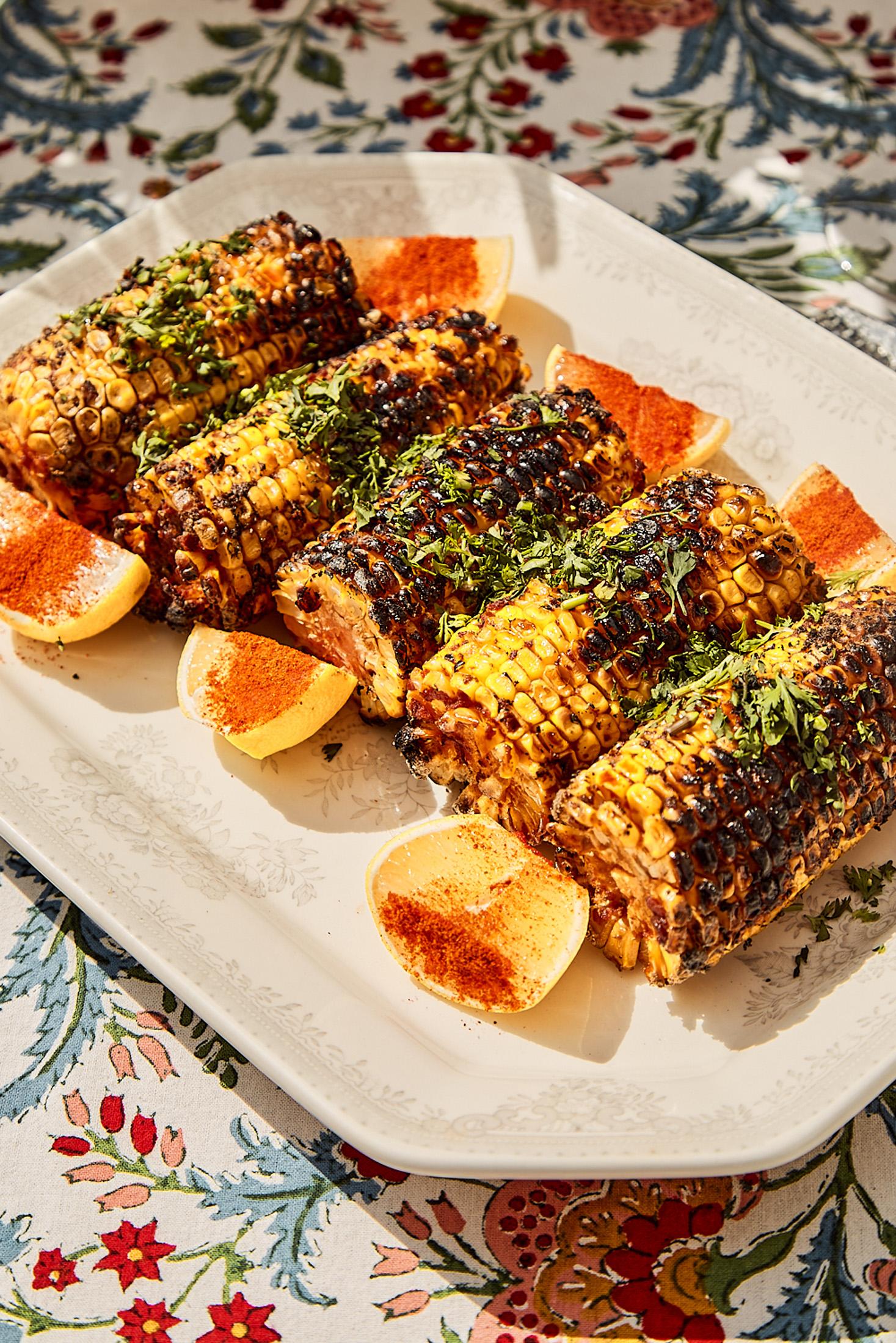 A plate features grilled corn on the cob, garnished with herbs and accompanied by orange slices.