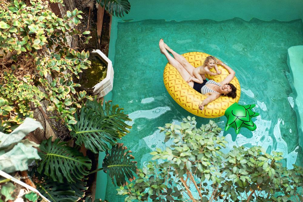 A woman and a child float on a yellow pineapple-shaped inflatable in a pool, surrounded by lush greenery. The woman's face is relaxed, while the child’s face is turned away,.