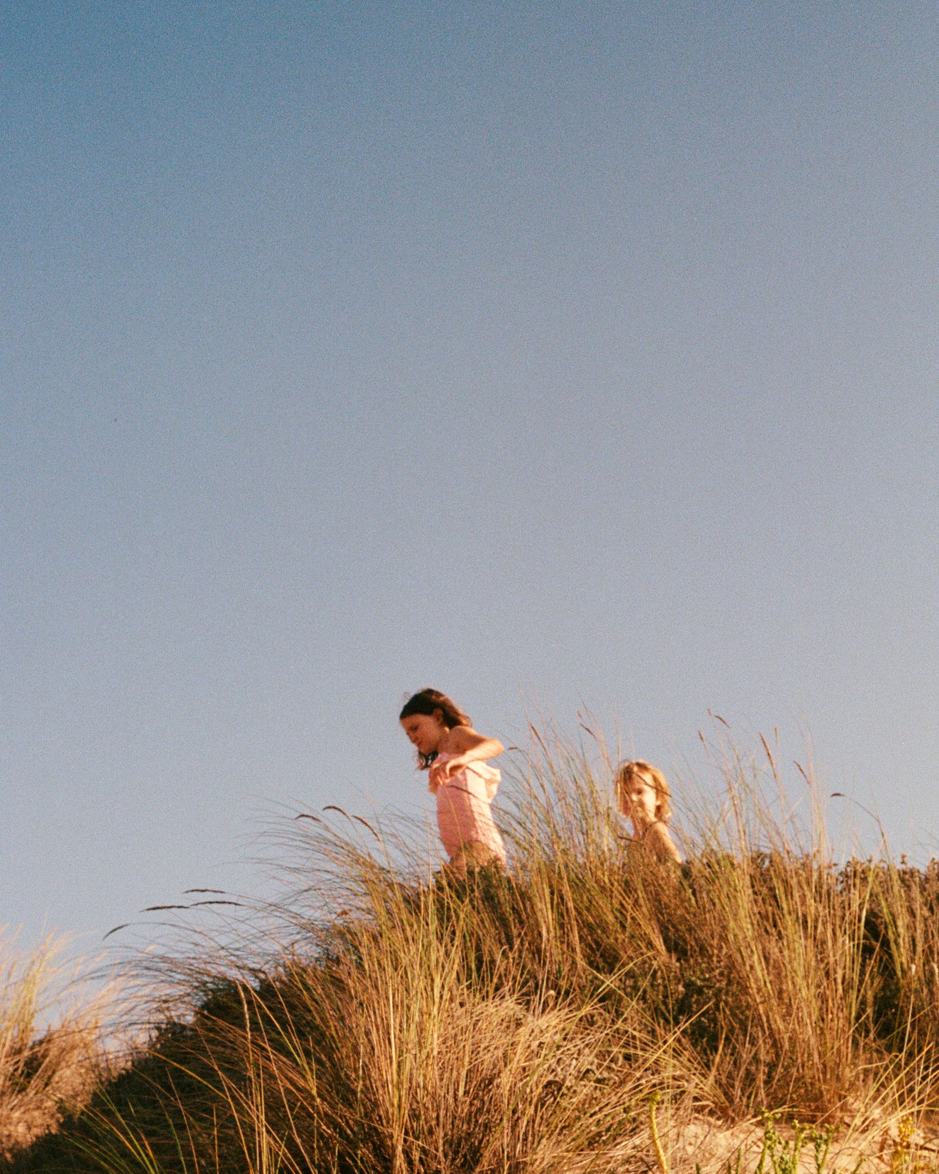 Two young girls playing in a sand dune, one wearing a pink dress and the other in a yellow sundress, both surrounded by tall grasses and wildflowers.