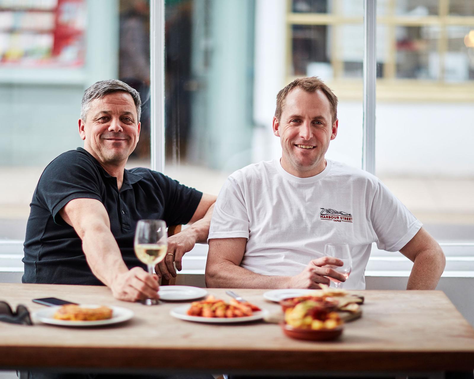 Two men sitting at a table, smiling and holding wine glasses, with a city street visible through the window behind them.