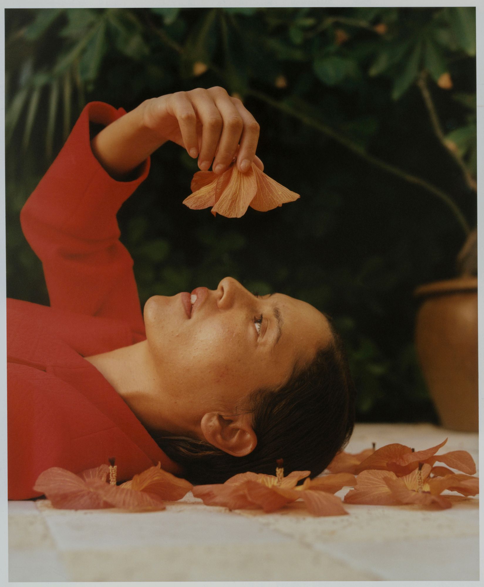 A woman in a red jacket lies on a bed of flowers, surrounded by petals and flowers in her hand.