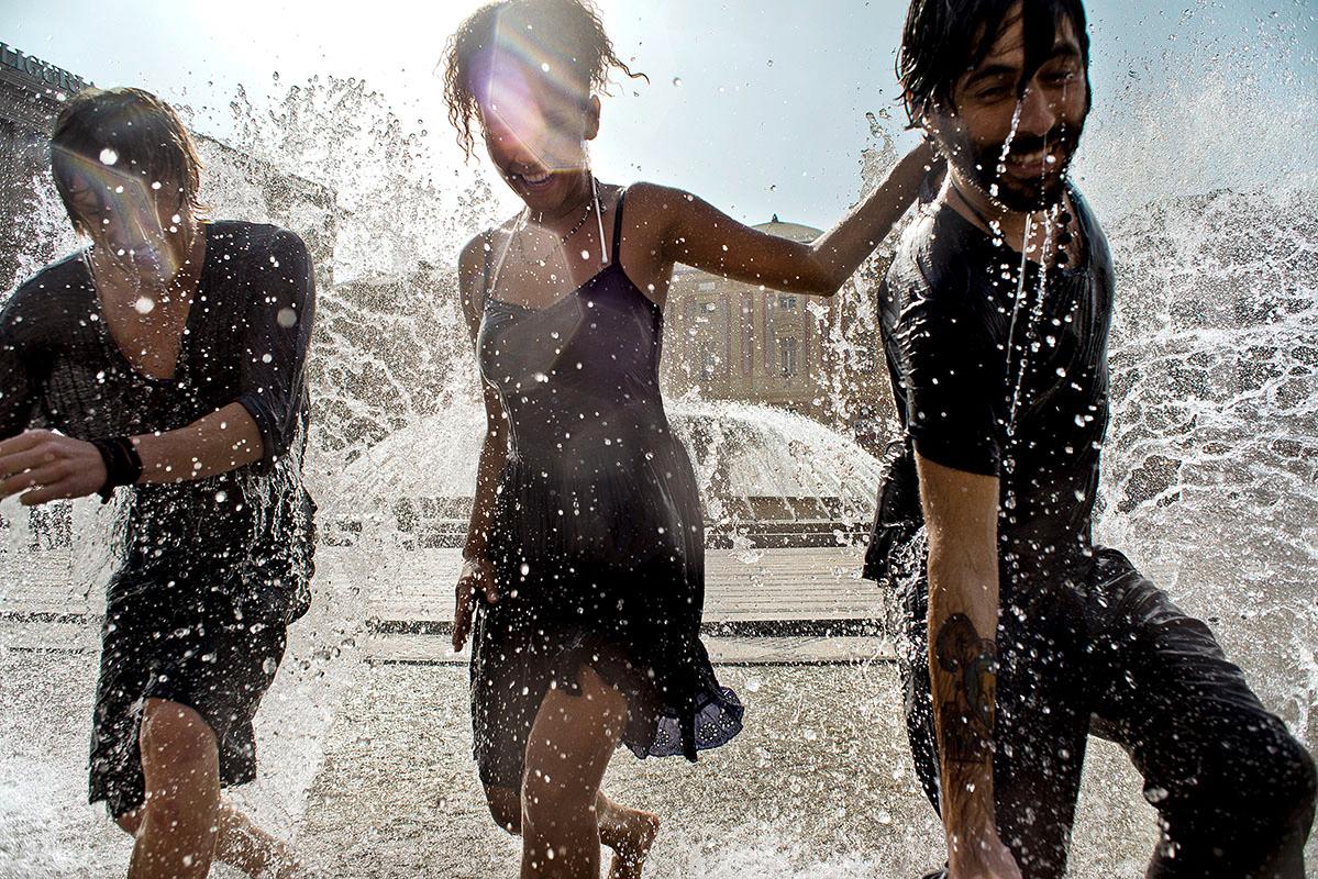 Three people, two women and one man, are running through a large fountain in the city, wearing black clothing.