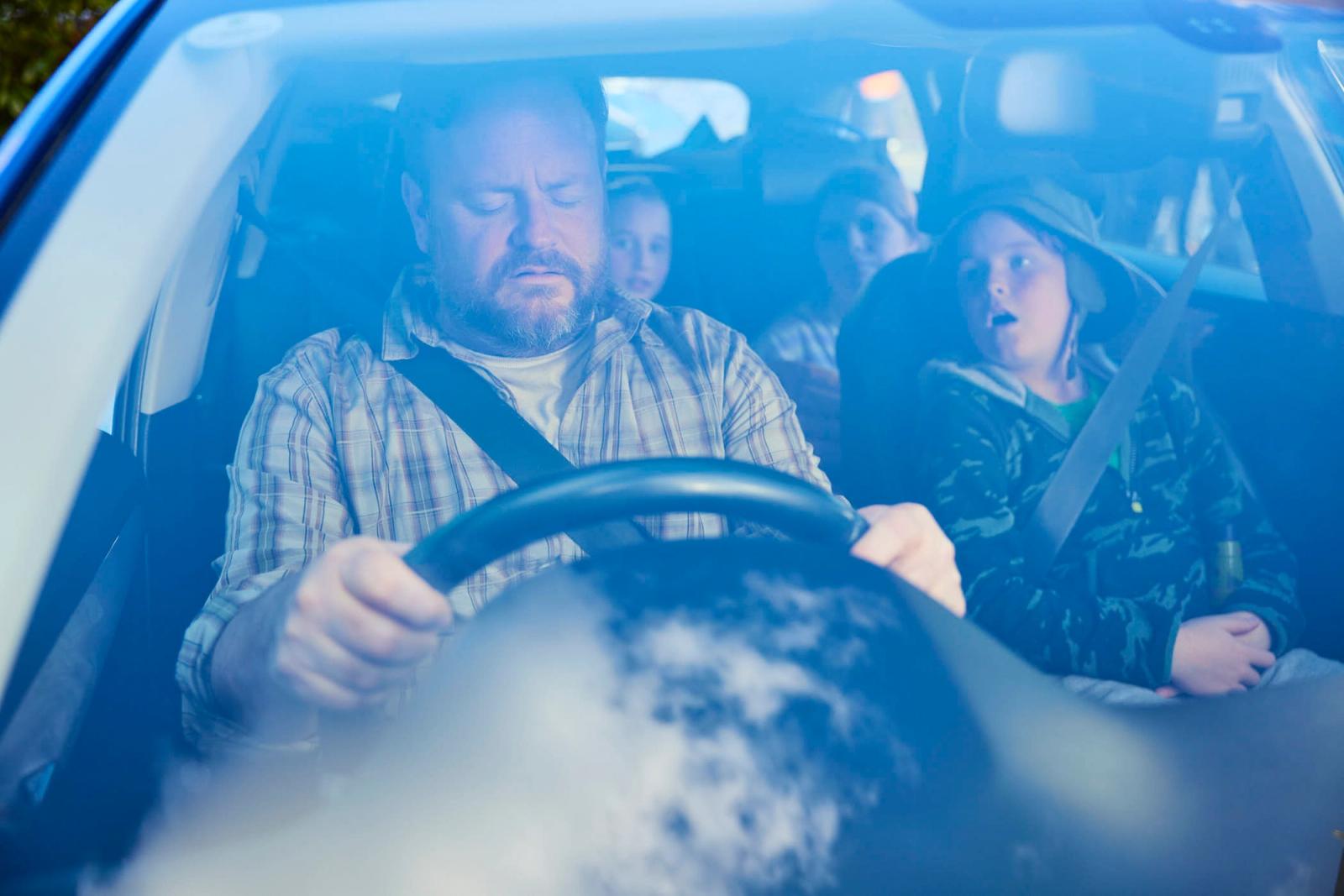 A man with a beard and a plaid shirt is driving a car with a child in the front passenger seat. The child is wearing a green jacket and a hat, and appears to be looking at.