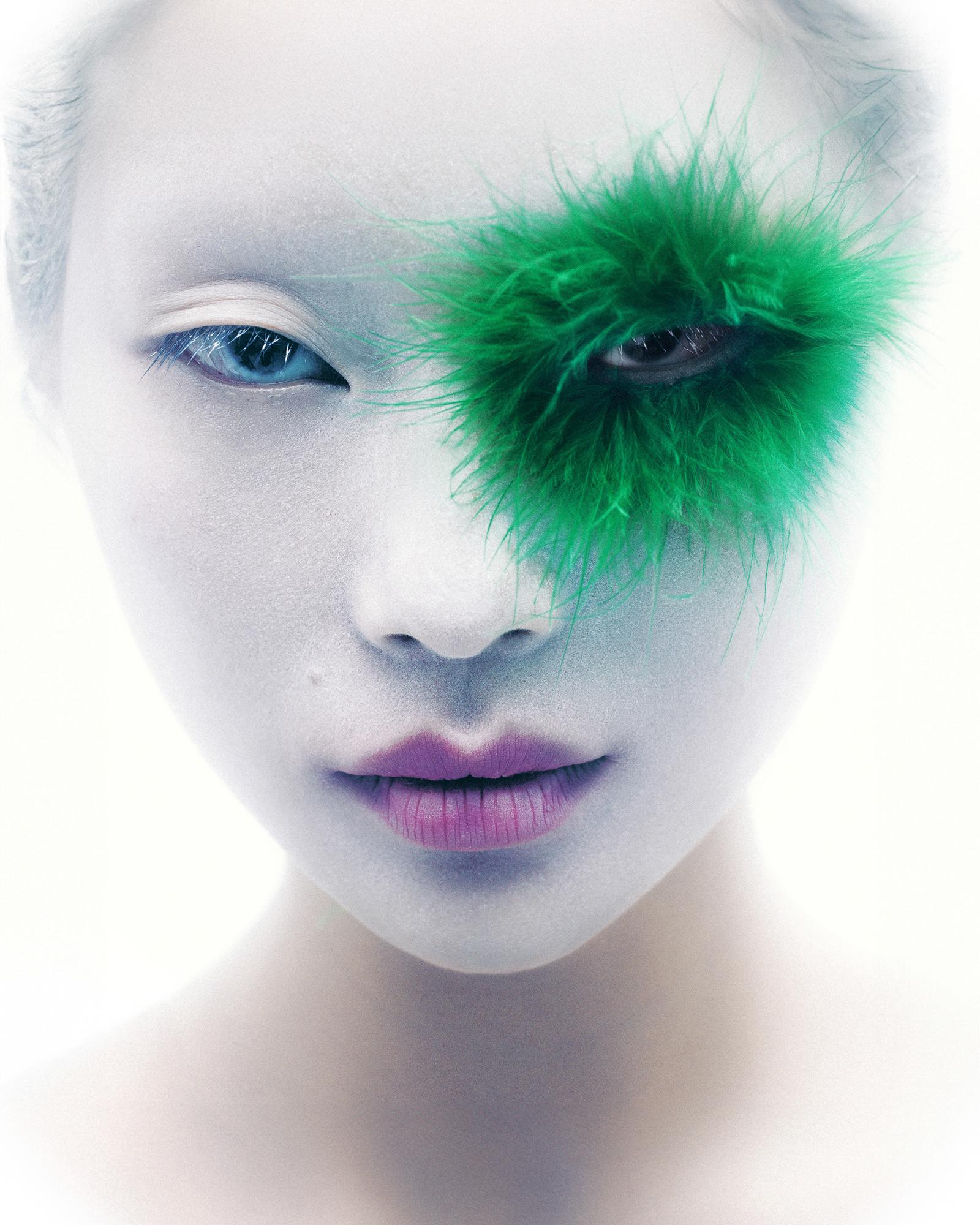 A close-up of a woman's face with a green, fuzzy, eye-like makeup design on her left eye, covering her eyelid and extending to her cheek.