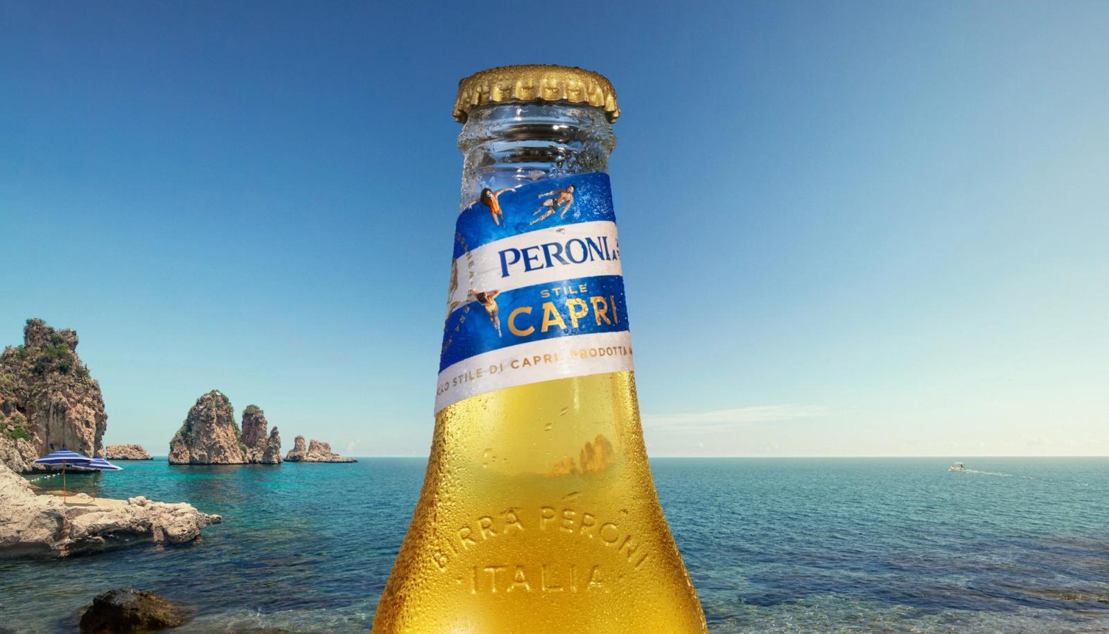 A bottle of Peroni beer on a rocky beach with a clear blue sky and a few rocky outcrops in the distance.