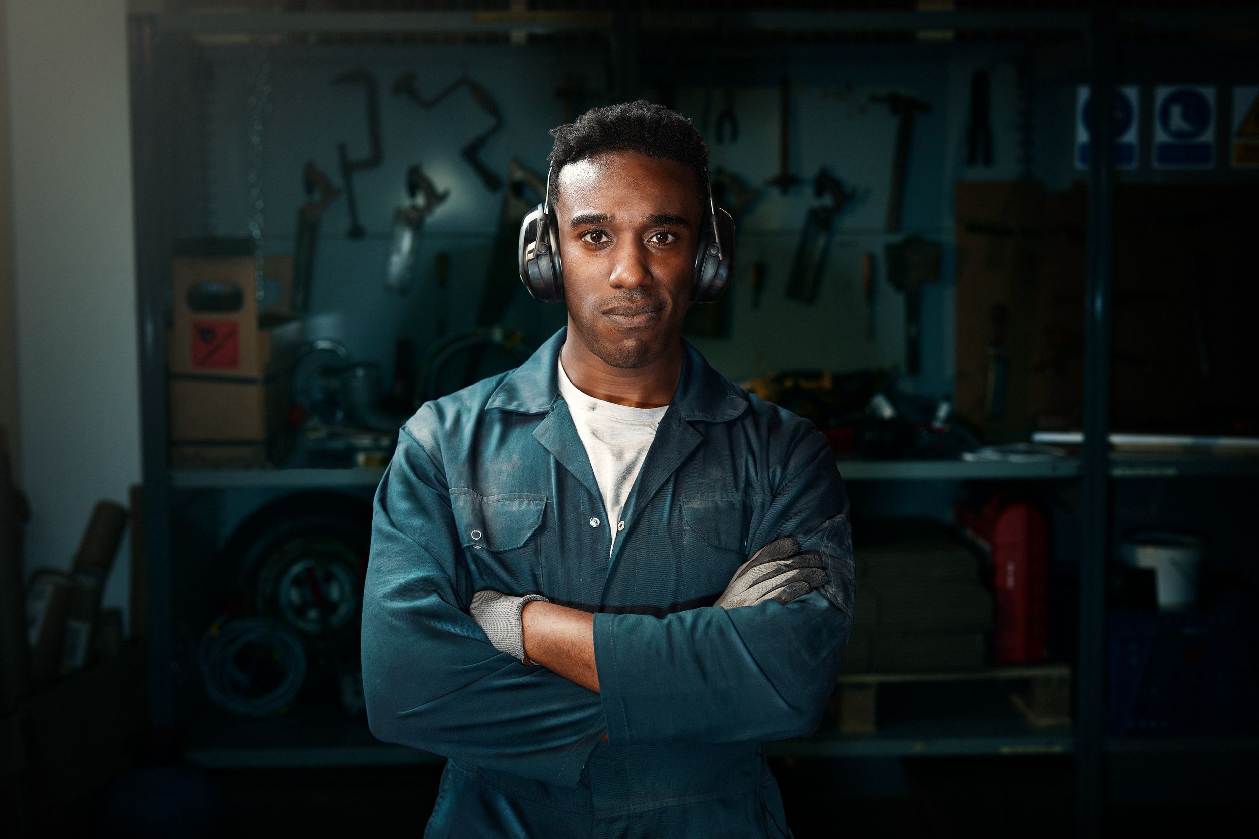 A young man in a blue jumpsuit and white t-shirt stands in a workshop surrounded by tools and machinery.