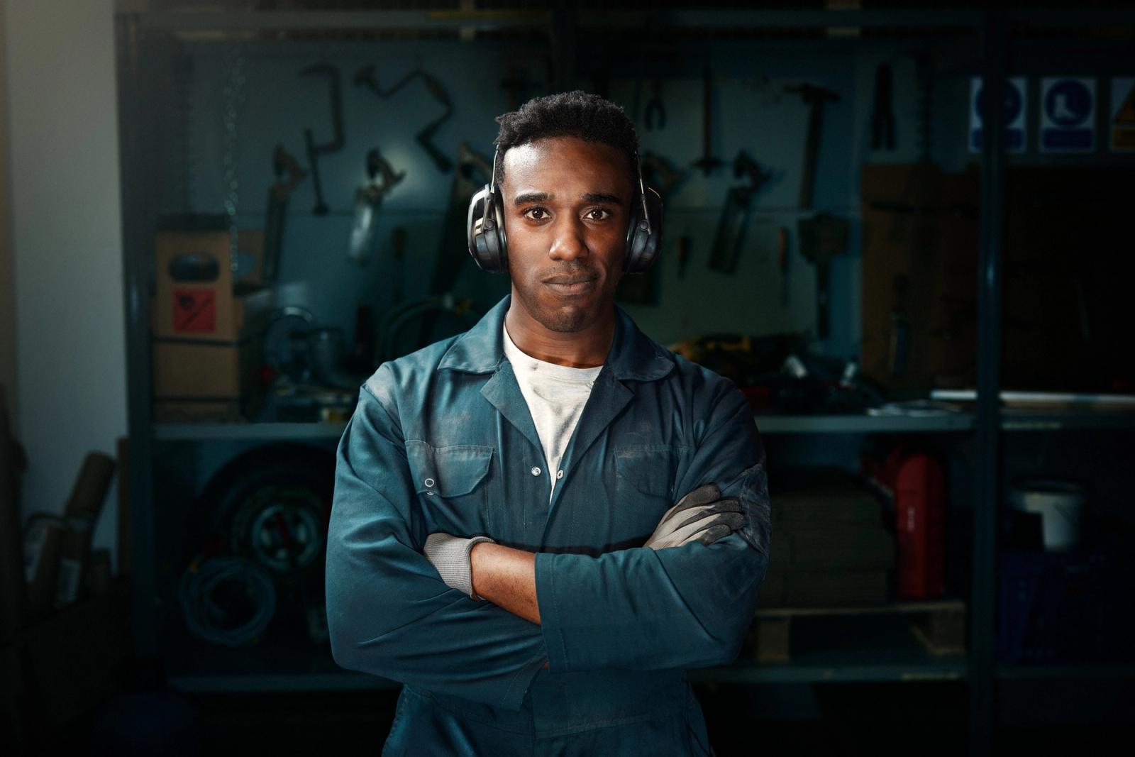A young man in a blue jumpsuit and white t-shirt stands in a workshop surrounded by tools and machinery.
