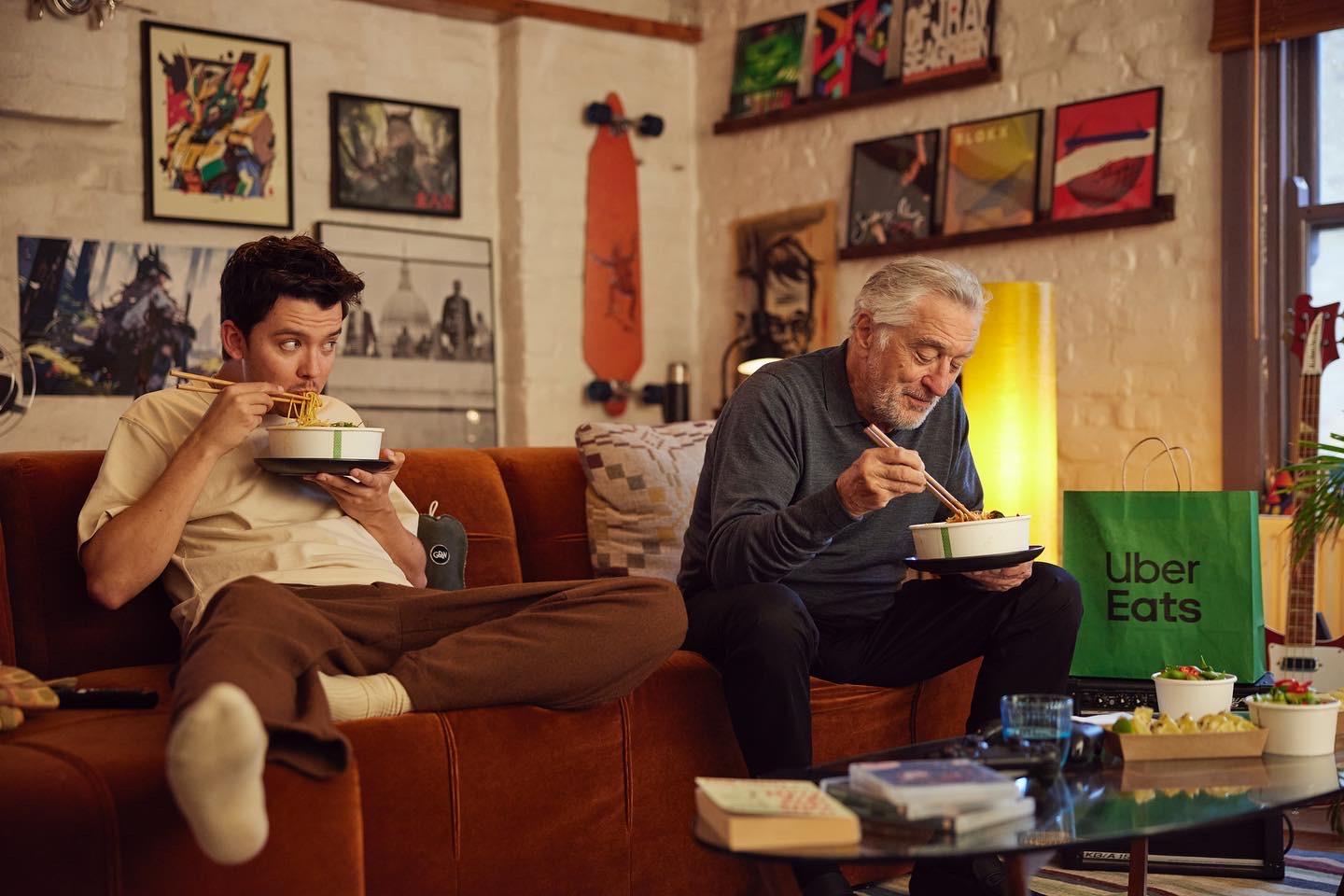 Two men sitting on a couch eating noodles from bowls with a TV in the background.