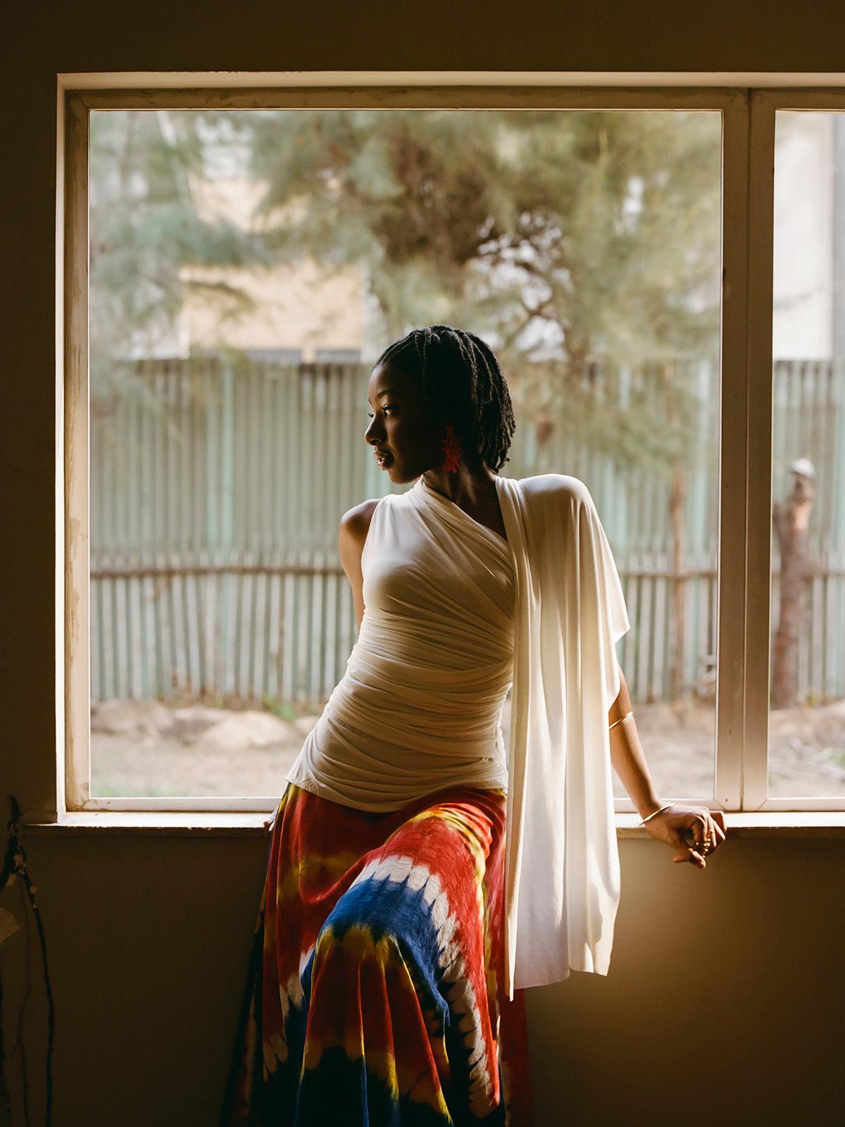 A woman with dreadlocks and a white top, standing in front of a window with a blurred background, wearing a colorful, long skirt with a red, blue, and yellow pattern.