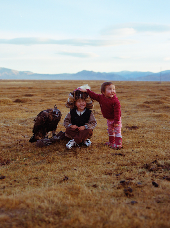A young boy and girl in traditional Mongolian clothing sit in a field with an eagle on a rock and mountains in the background.