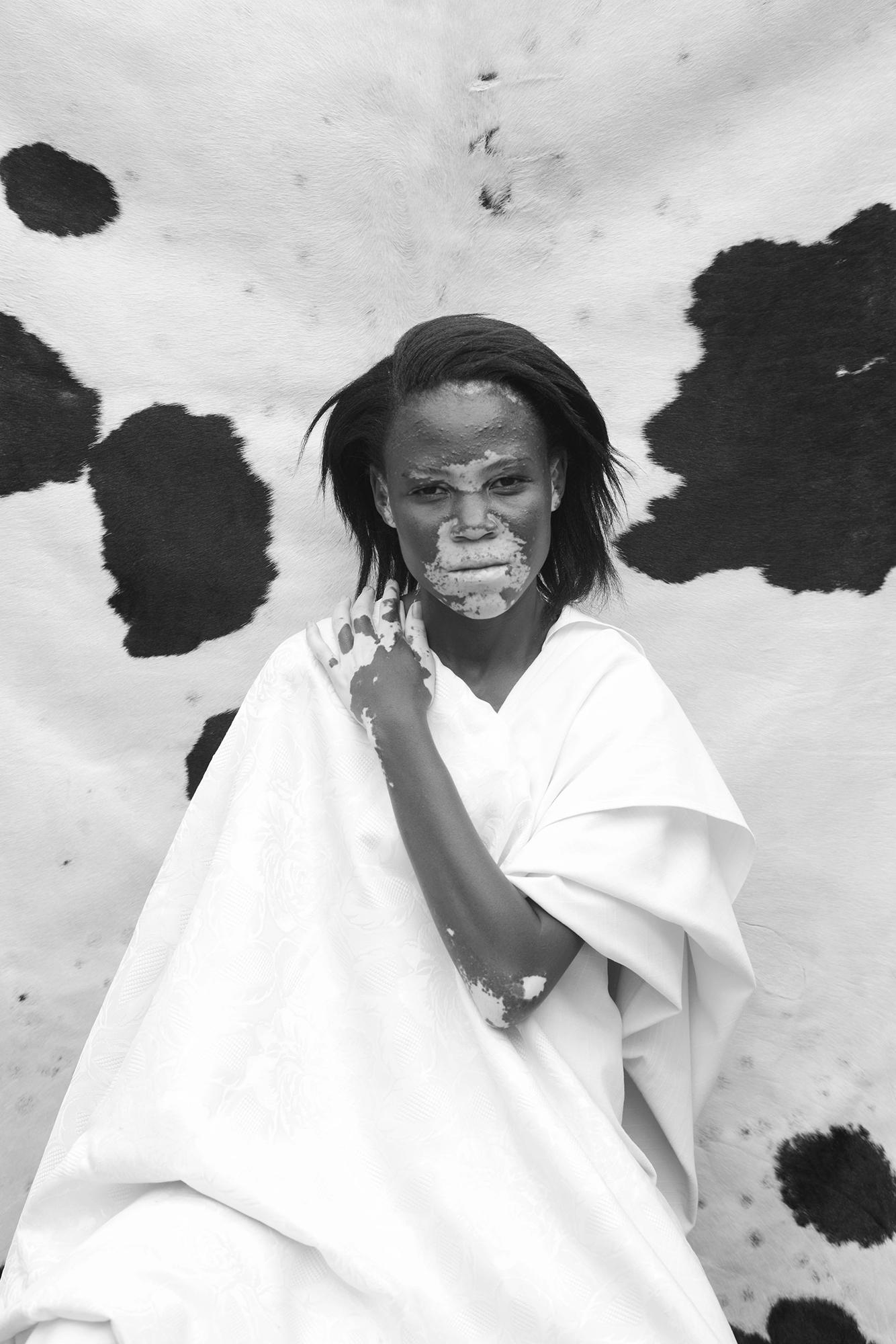 A woman with a white face and a white dress, standing in front of a white background with a few black spots.