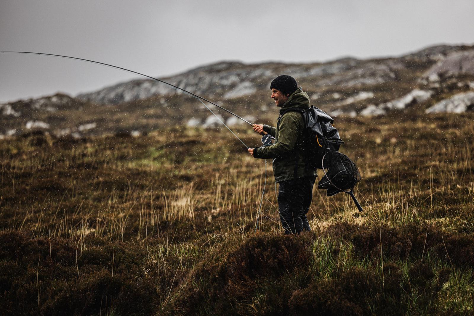 A man in a green jacket and black hat is standing in a rugged, mountainous landscape, holding a fishing rod and casting a line into a nearby stream.