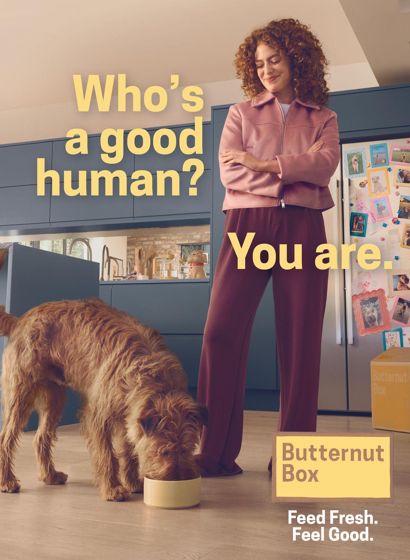 A woman in a pink jacket and purple pants standing in a kitchen, with a dog eating from a yellow bowl on the floor.