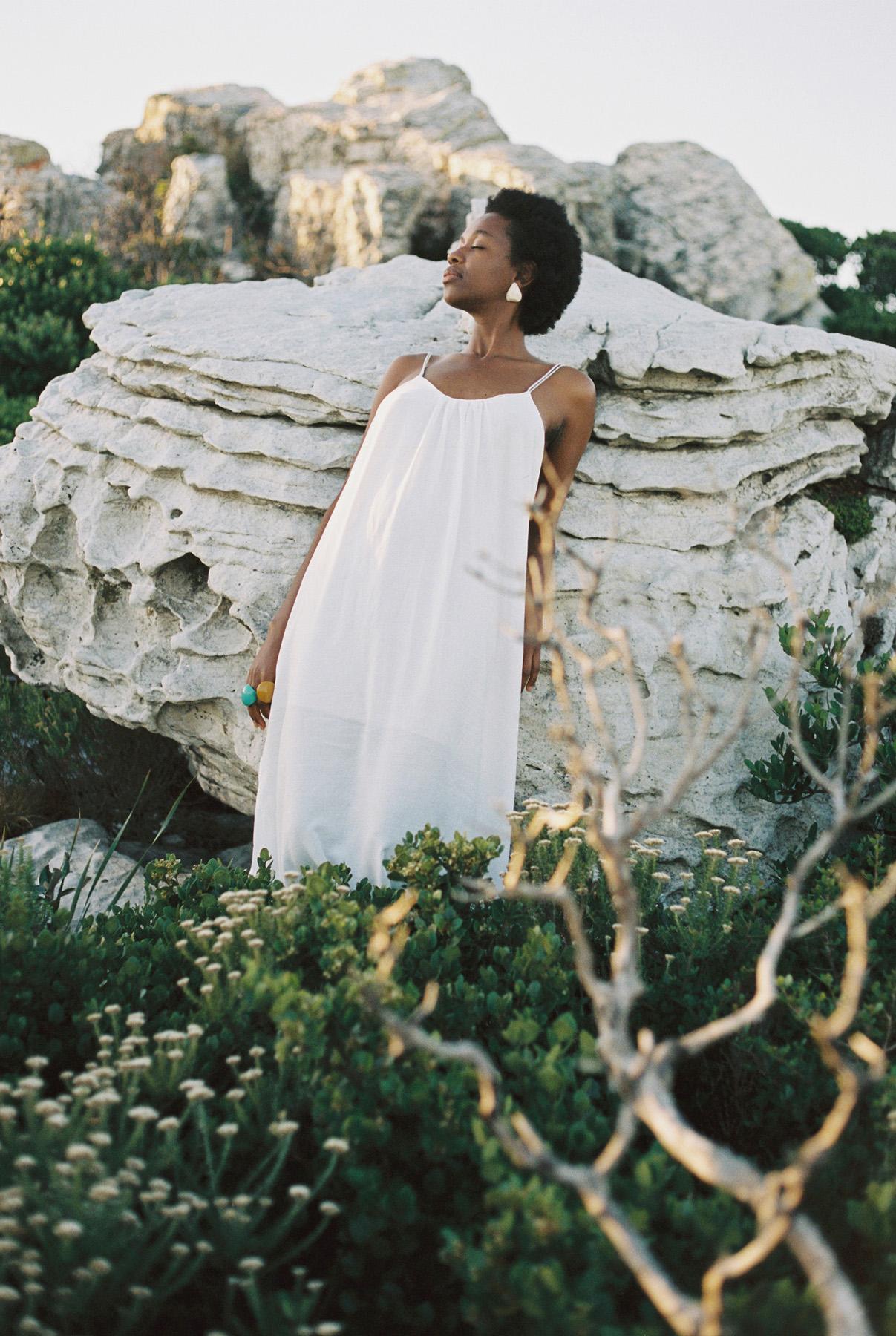 A woman in a white dress stands in a rocky, desert-like landscape, surrounded by greenery and wildflowers.