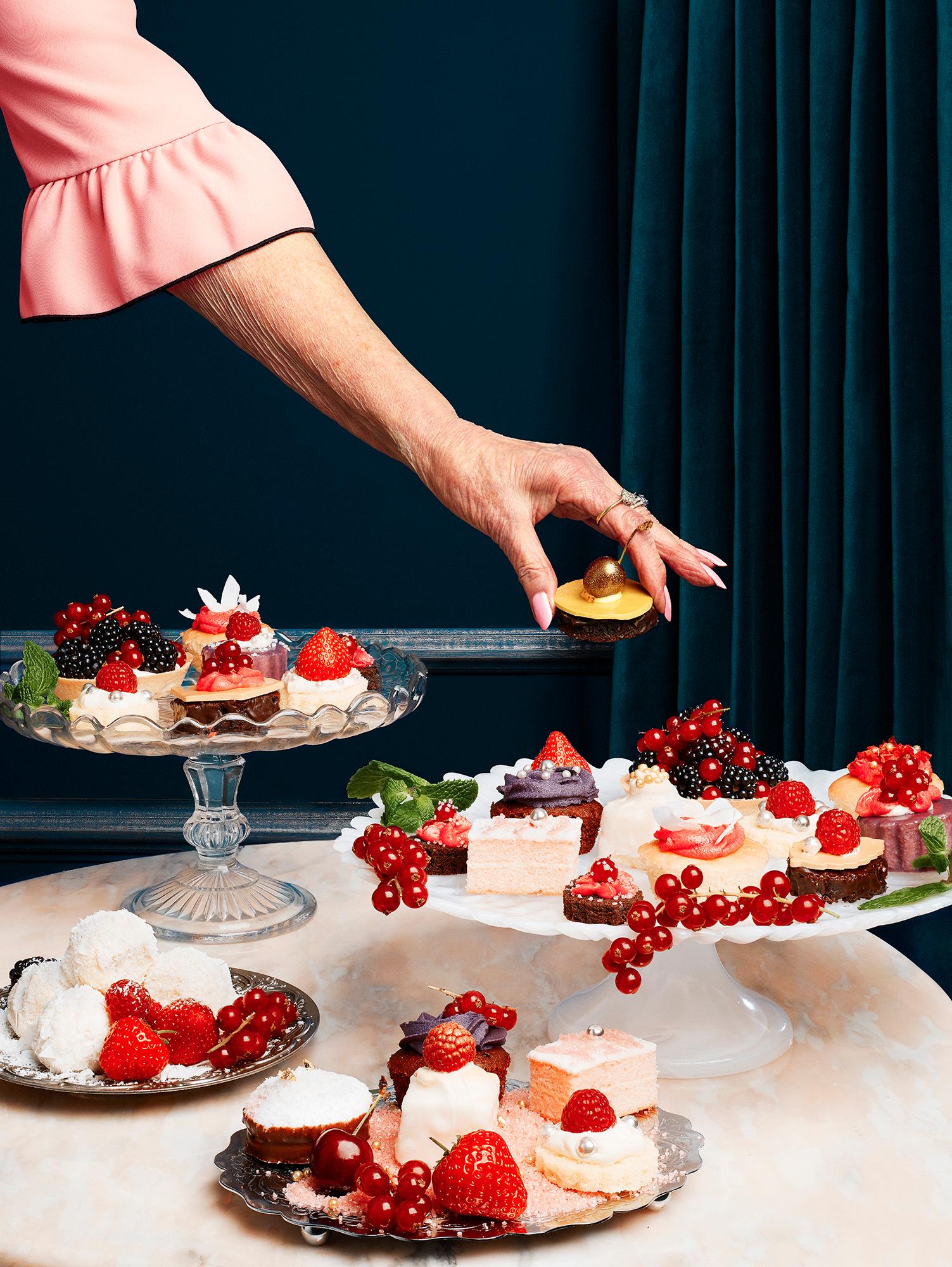 A woman's hand reaching for a chocolate cupcake with a gold ball on top, surrounded by various desserts and fruit on a marble table in front of a dark blue wall.