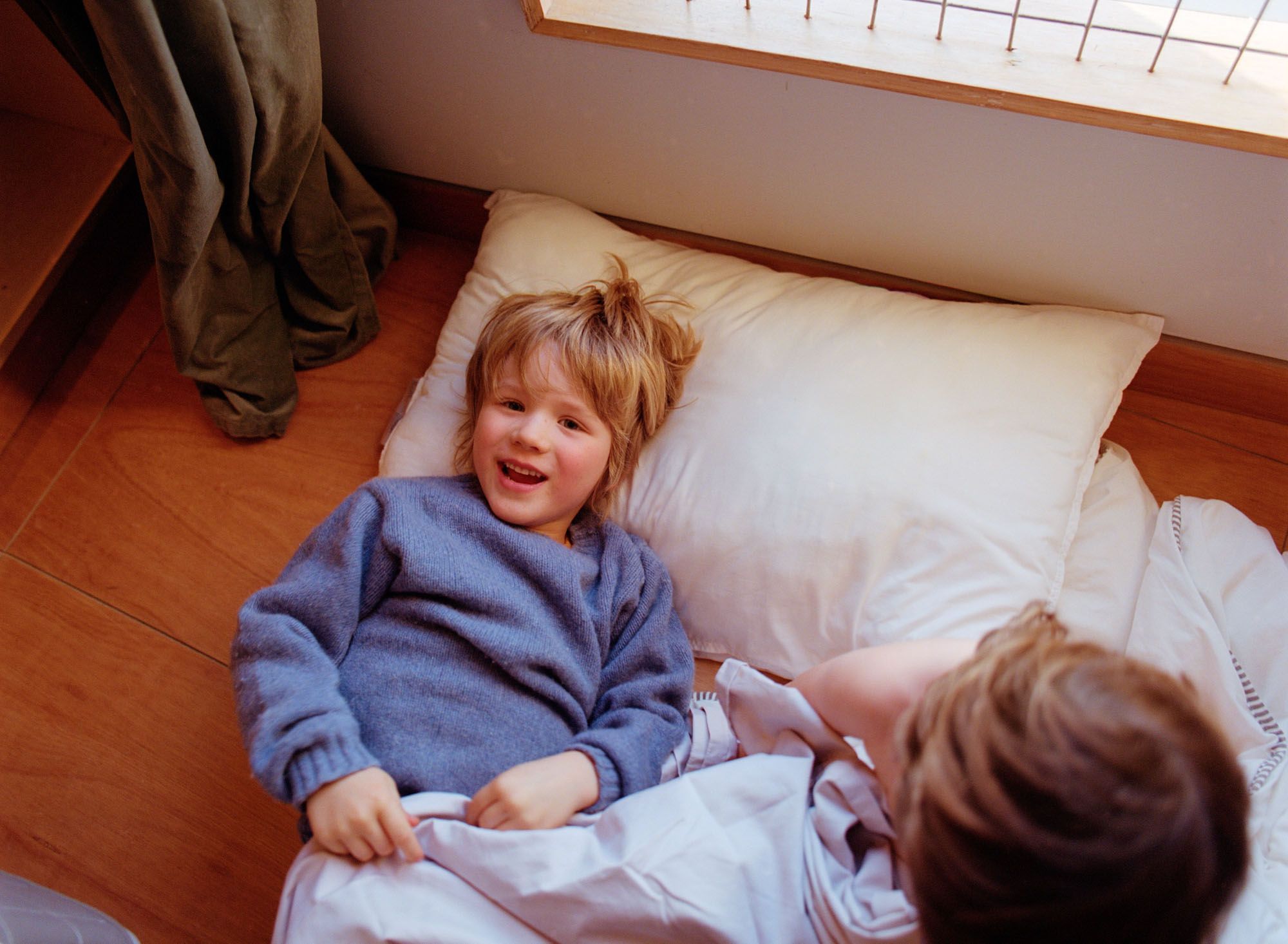 Two young children, likely siblings, are lying on a floor, playing together, with one child wearing a blue sweater and the other in a white shirt, both on a white blanket.