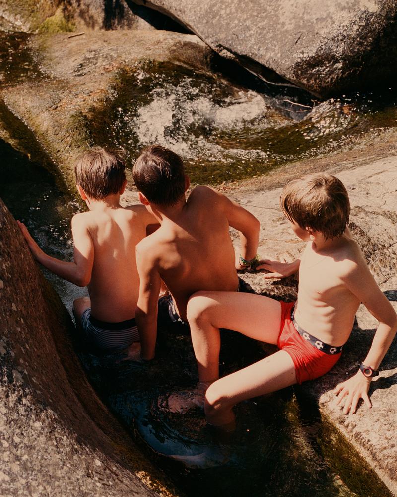 Three boys in swimsuits play in a rock pool surrounded by large boulders.