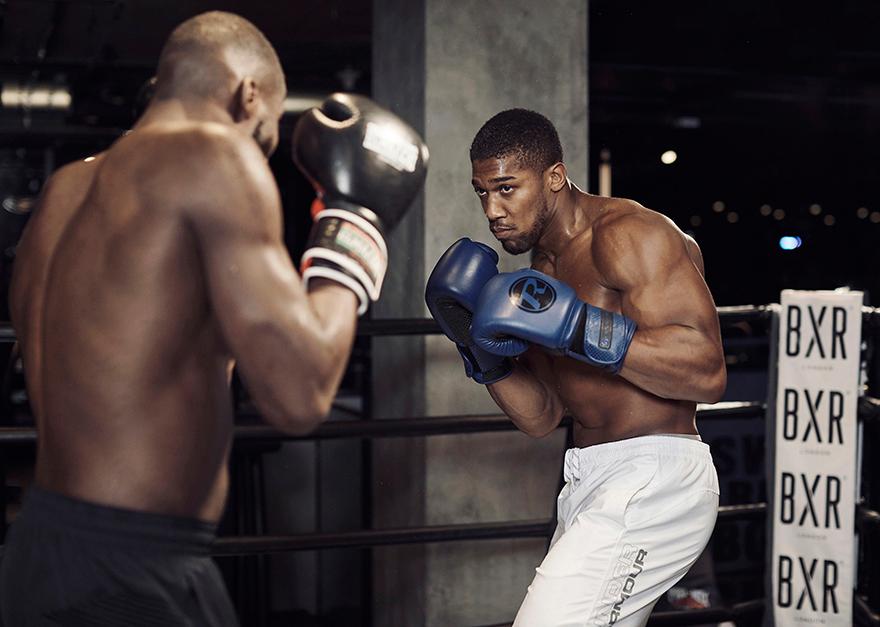 Two shirtless, muscular men in a boxing ring, one wearing blue boxing gloves and the other wearing black and white boxing gloves, both in a fighting stance.