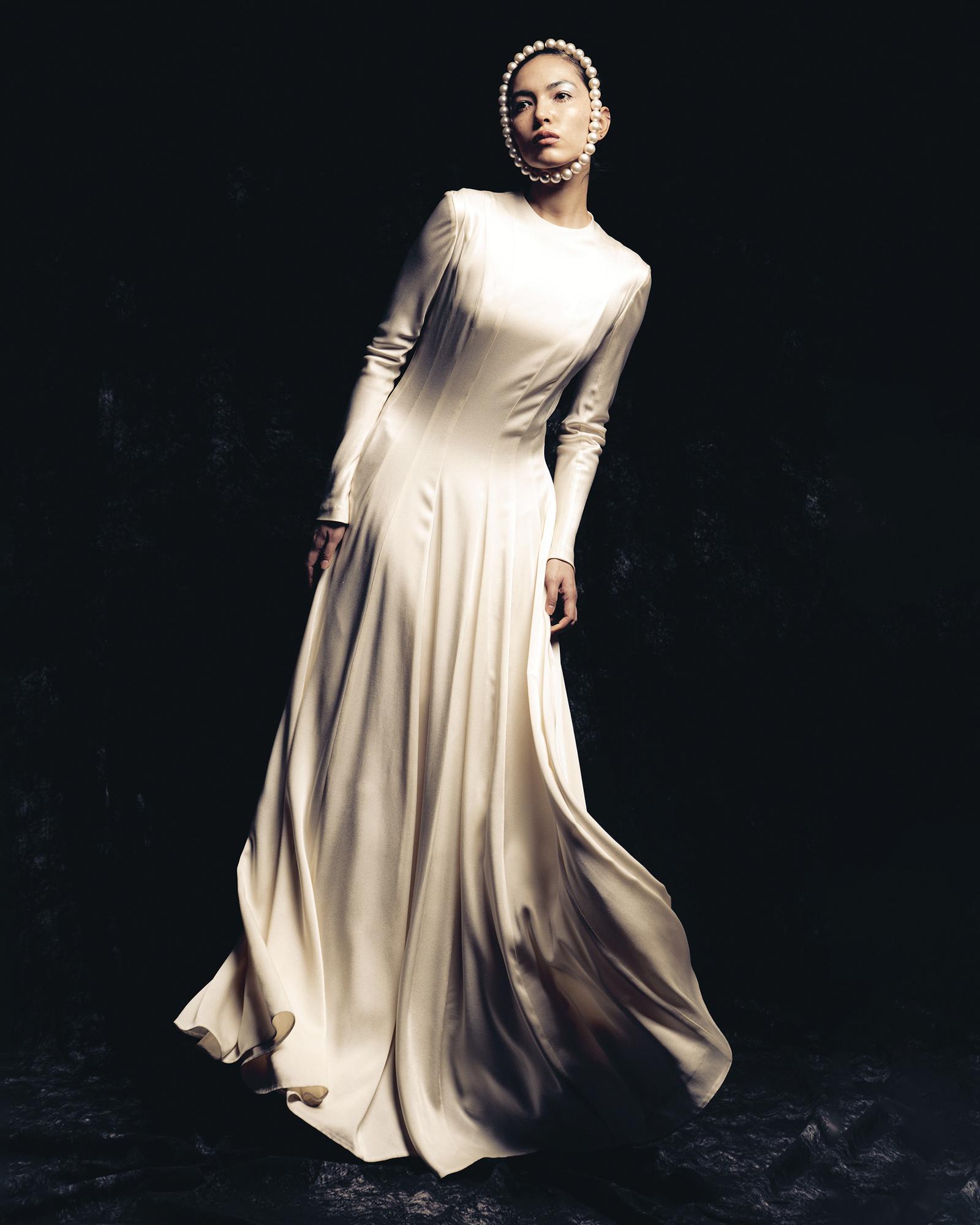 A woman in a white, long-sleeved, floor-length wedding dress with a high neckline and a full, flowing skirt, standing in a dark, dramatic setting.